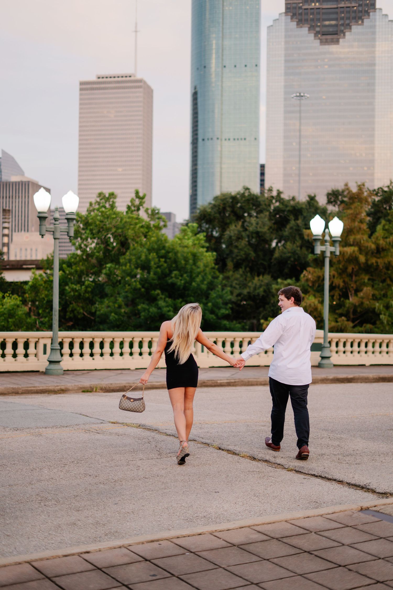 Couple holding hands walking toward city skyline at sunset, woman in black mini dress with handbag, man in white shirt.