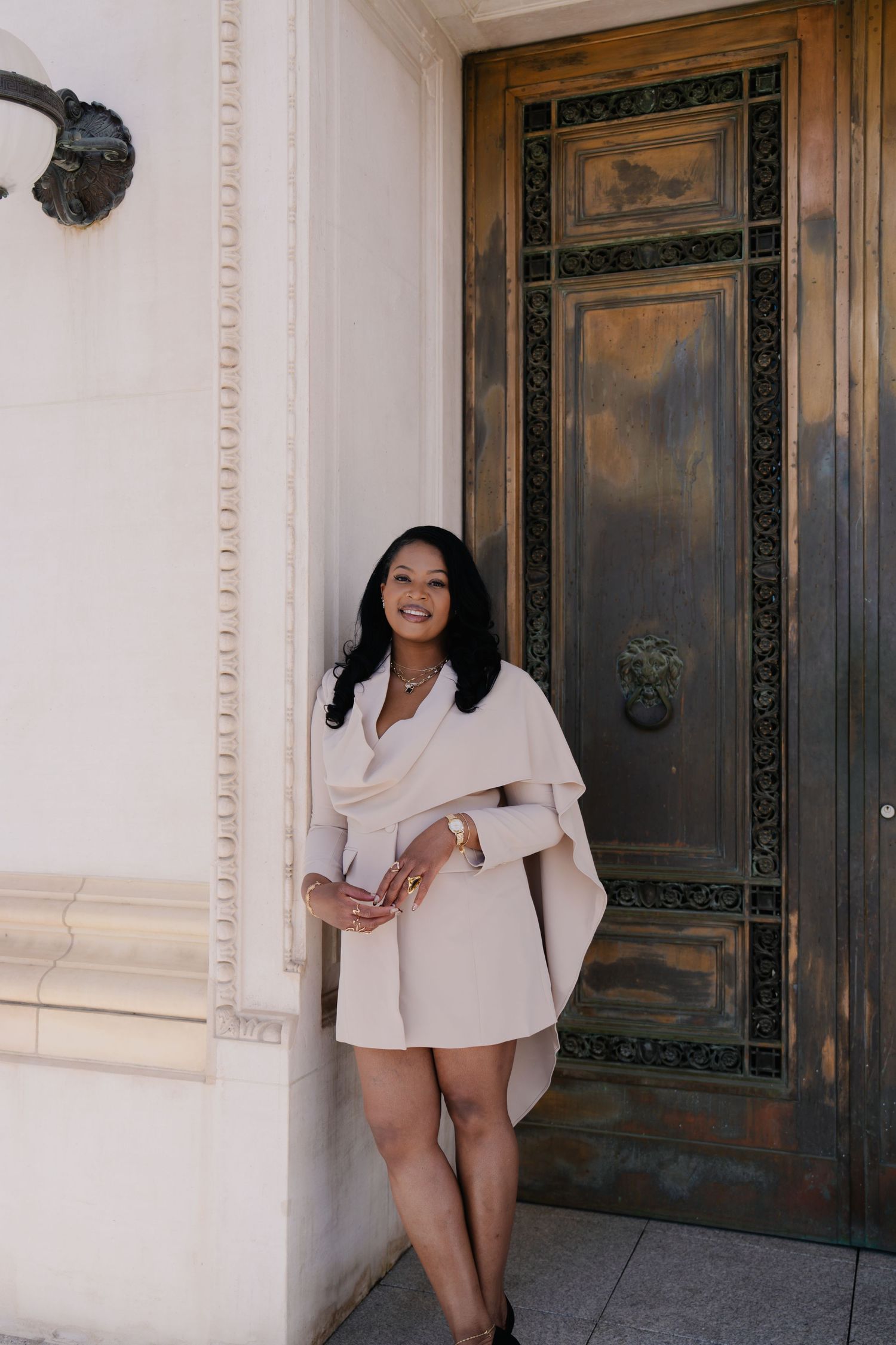 Woman in white cape dress smiling beside ornate bronze door and white column.