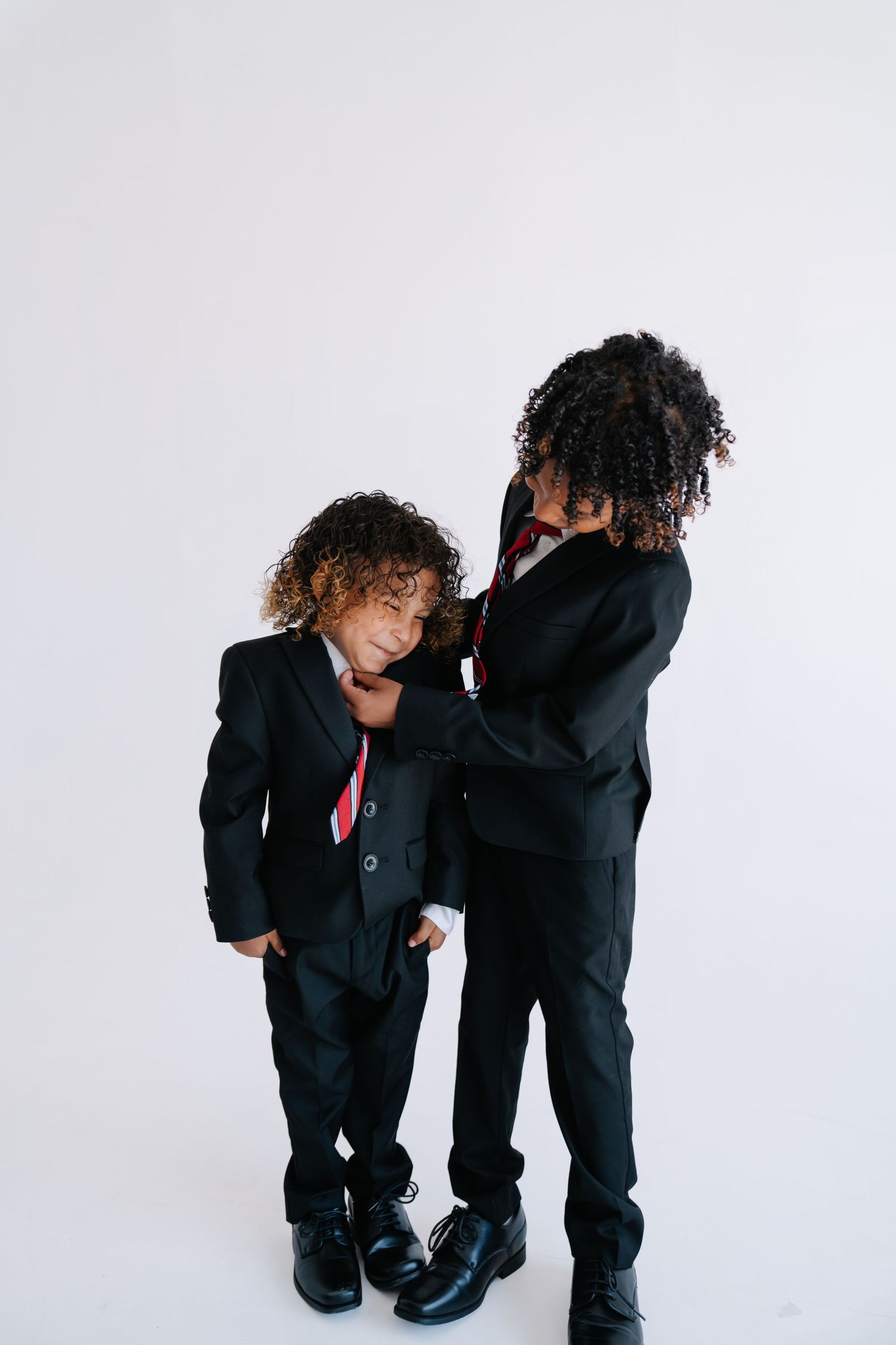 Two young boys in matching black suits and red ties pose together against a white background.
