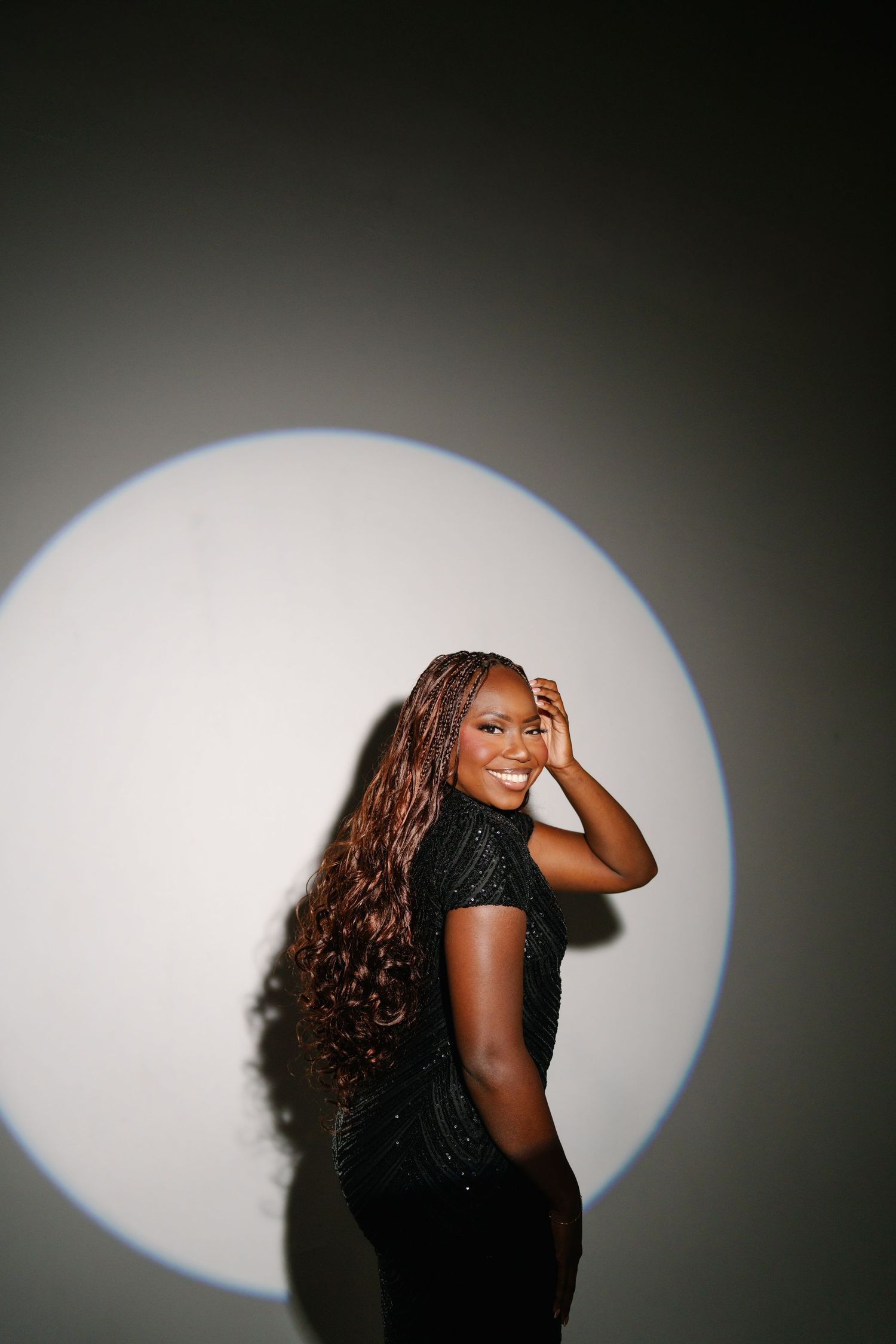 Smiling woman with long curly hair in black outfit poses against wall with circular spotlight backdrop.