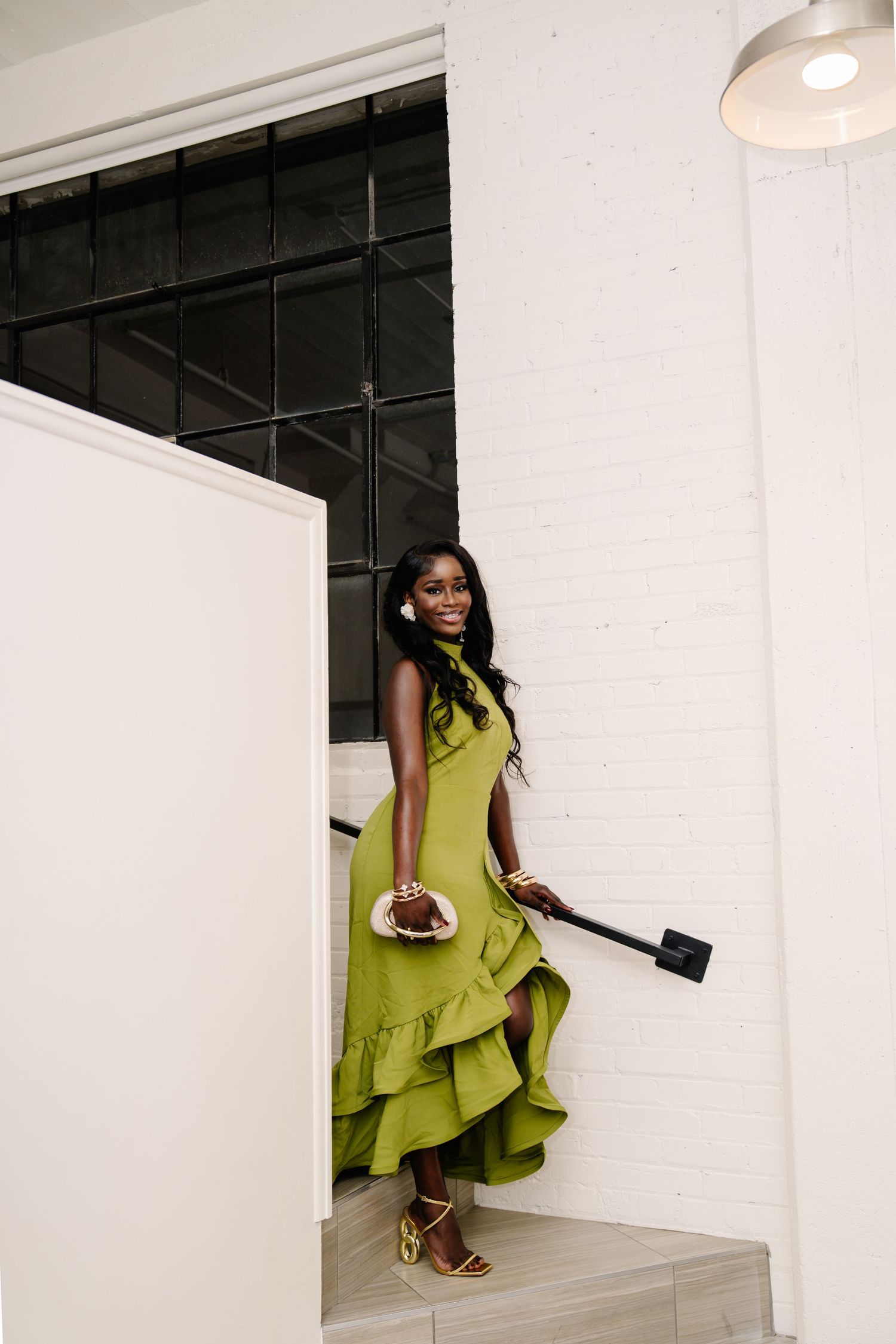 A woman in a flowing olive green ruffle dress poses elegantly on stairs, holding a clutch, wearing matching heeled sandals.