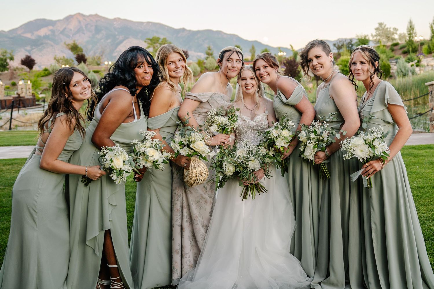 Bridesmaids in sage green dresses holding bouquets pose against mountain backdrop at sunset.