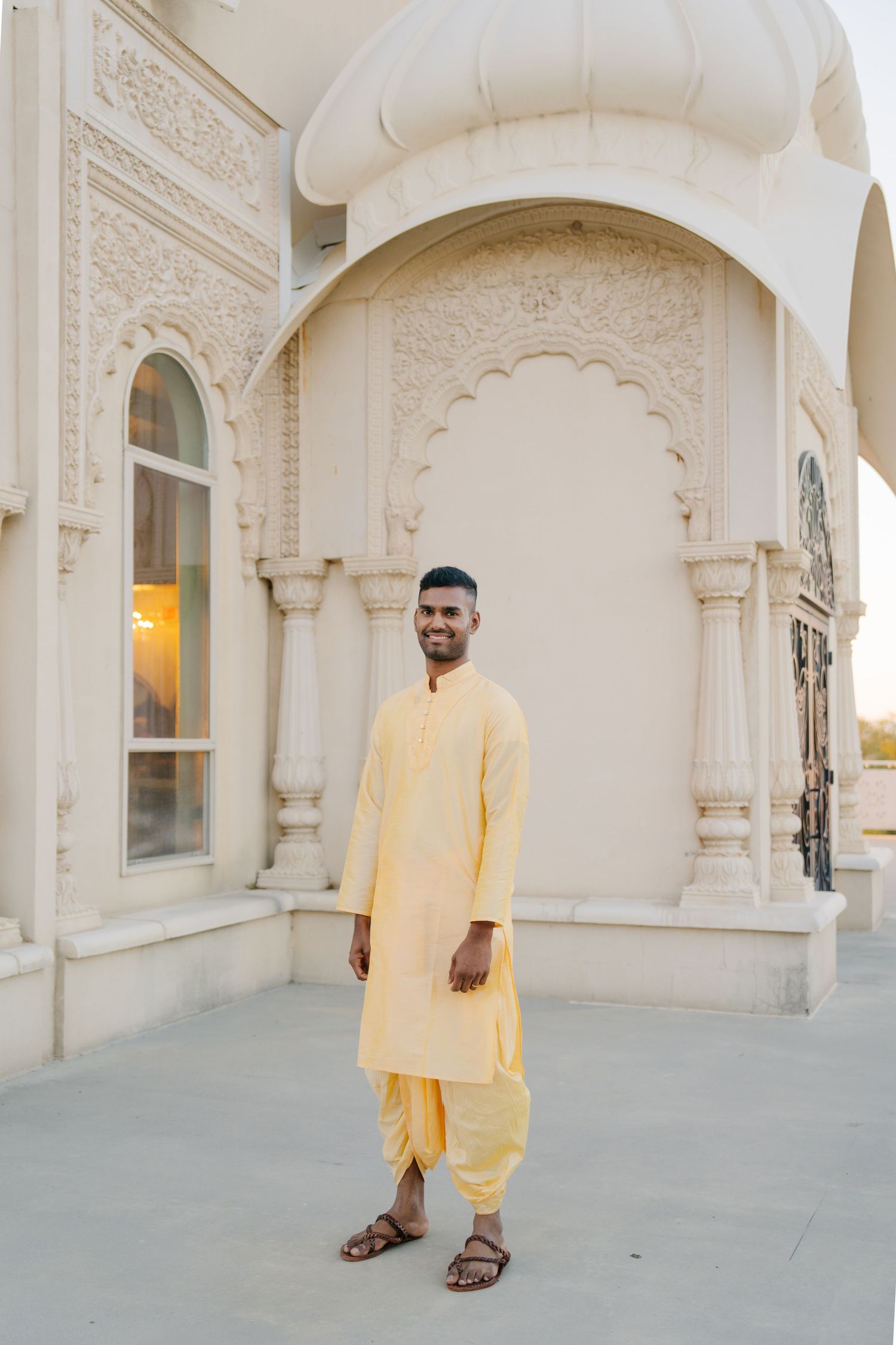 A person wearing a pale yellow kurta and dhoti stands in front of an ornate white temple facade.