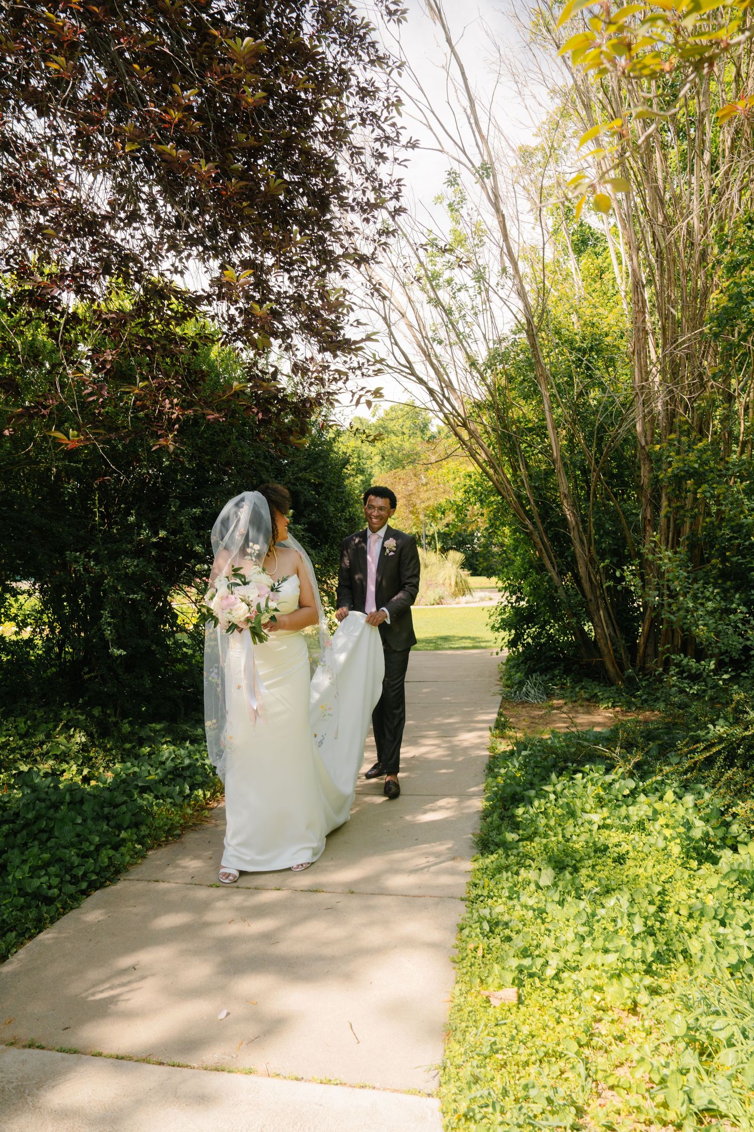 Bride and groom walking hand in hand along a sunlit garden path surrounded by lush green trees on their wedding day.
