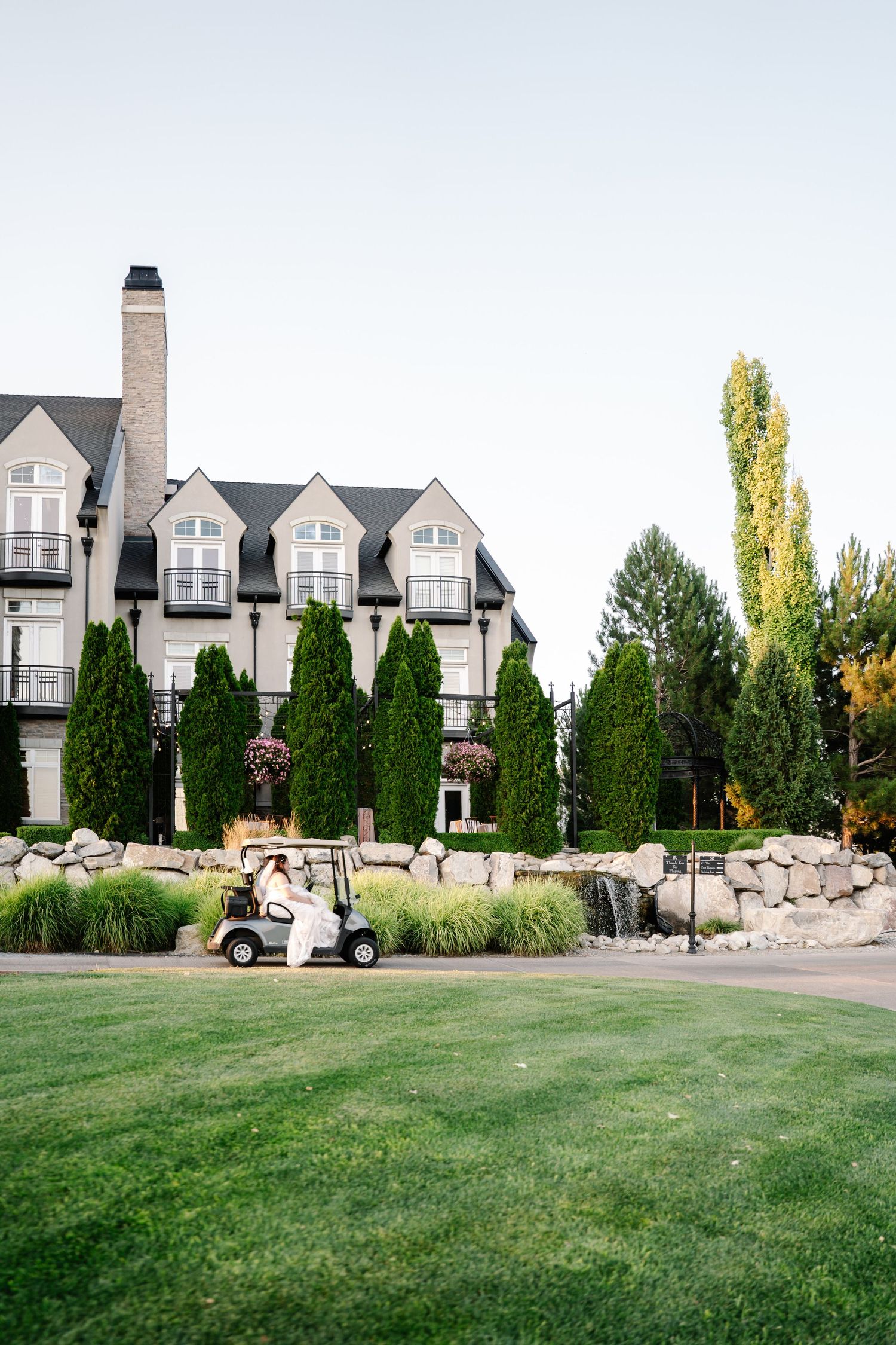A stately manor house with dormers sits on a manicured lawn with evergreen trees and a golf cart in the foreground.