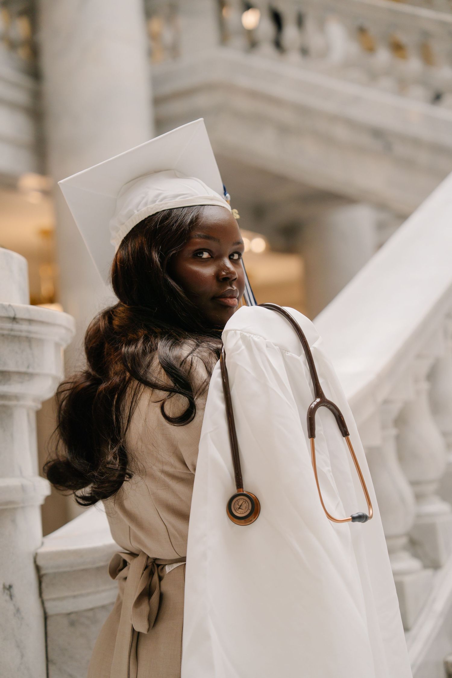 A person in a white headscarf and beige outfit poses against ornate white stone architecture.