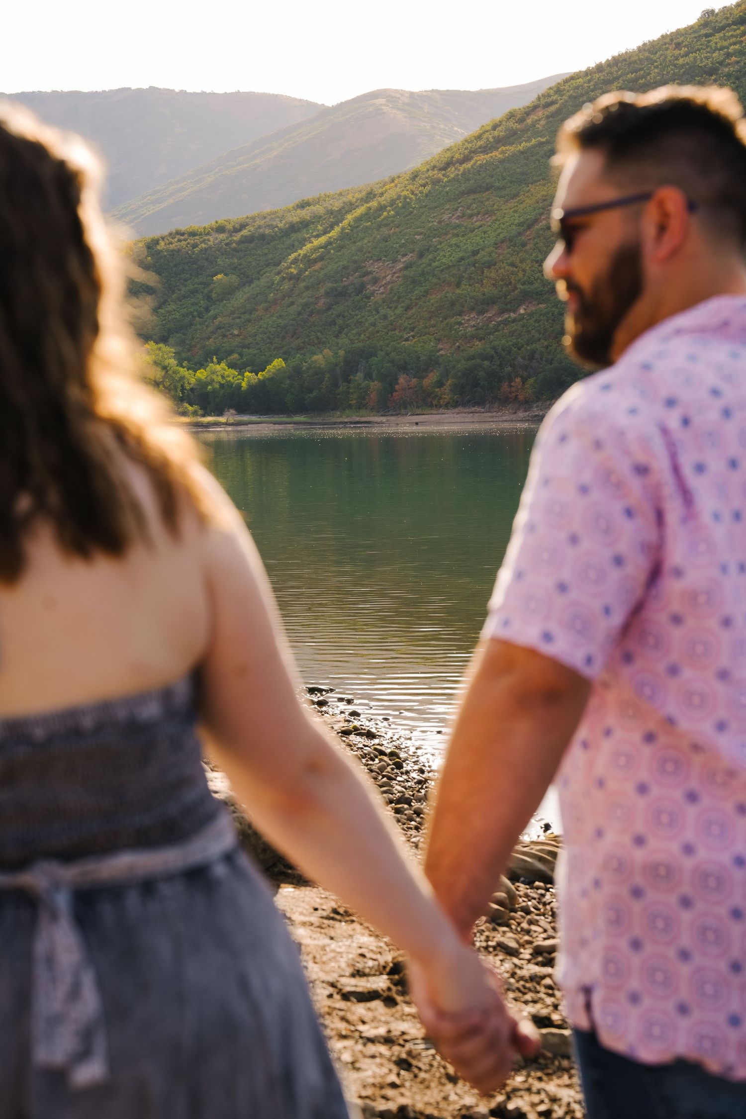 A couple walks along a mountain lake shore during golden hour.