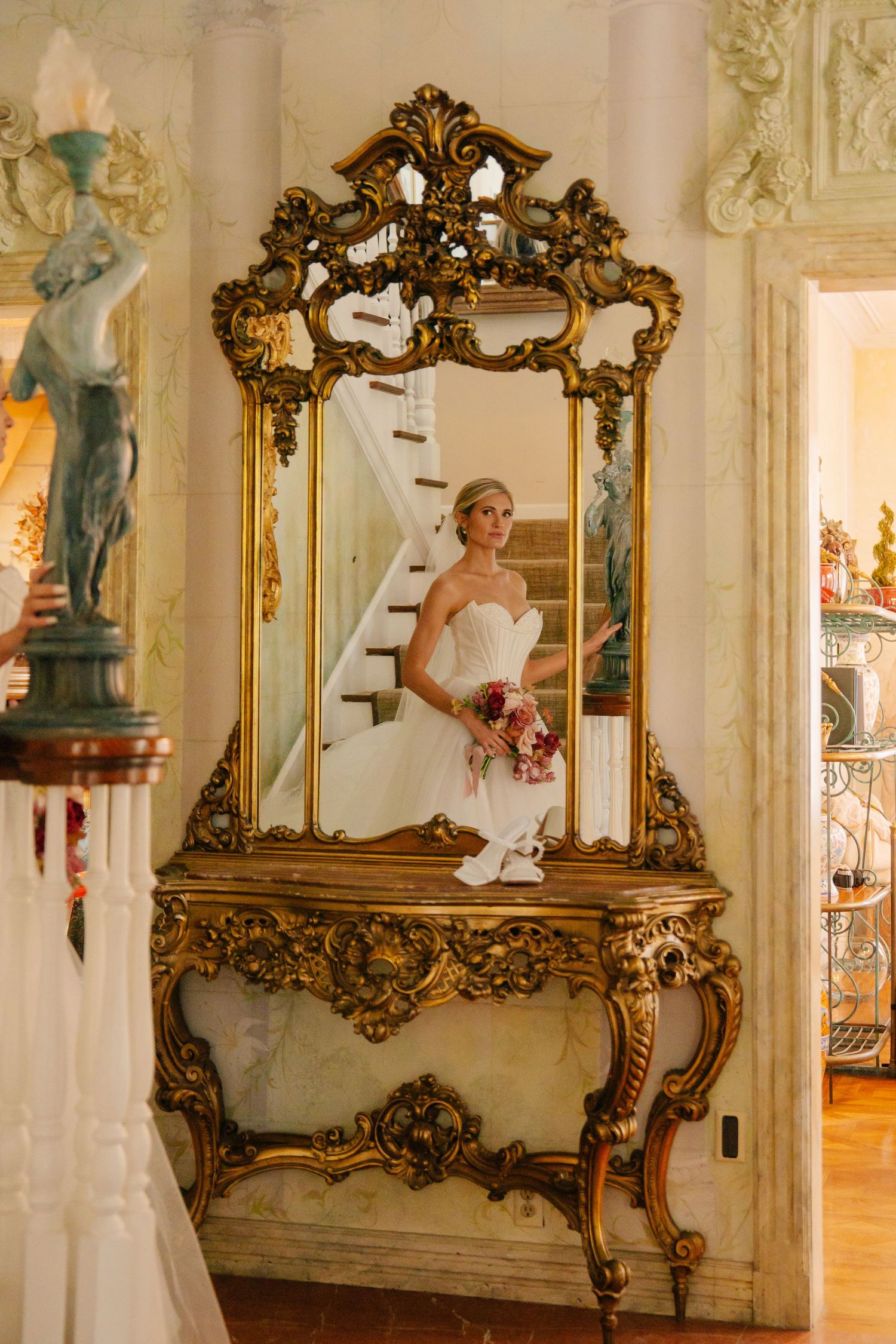 Bride in white gown reflected in an ornate gold mirror beside a decorative console table.