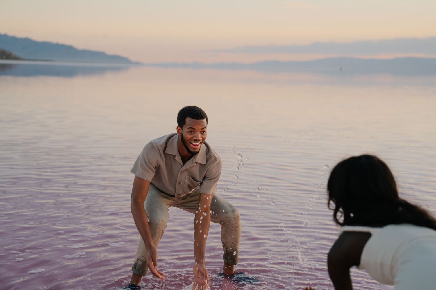 A playful couple enjoys a romantic moment in pink-tinted waters at dusk.