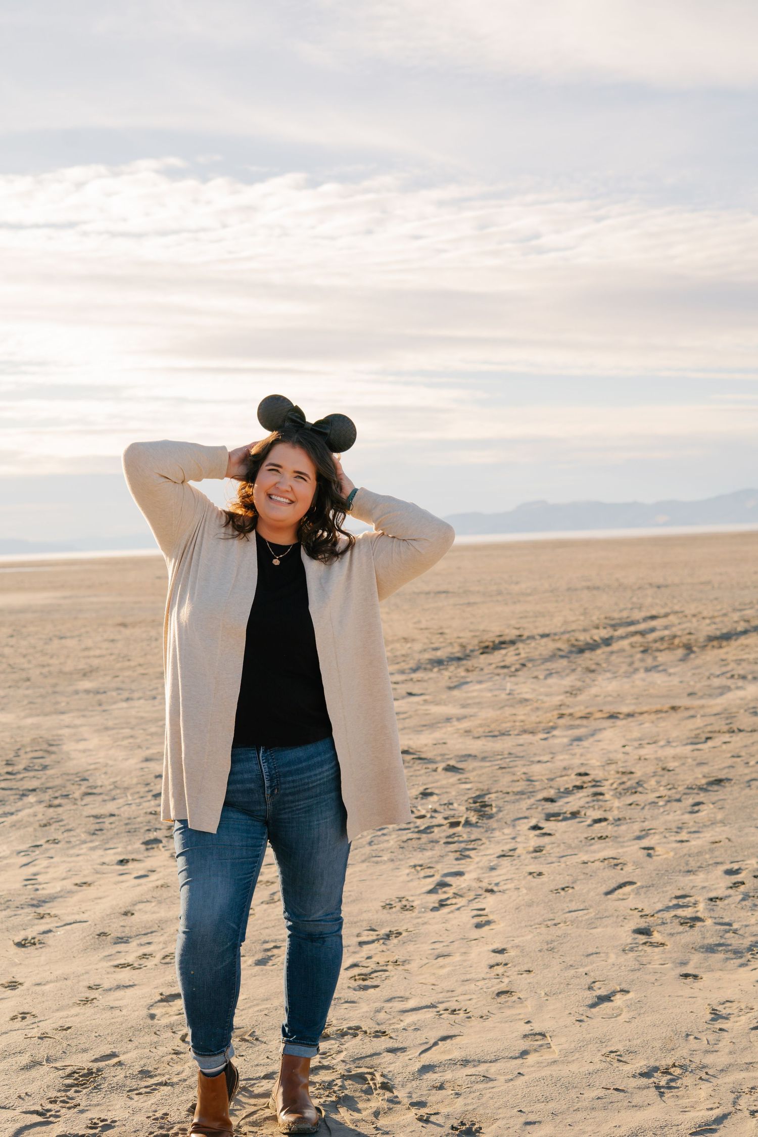 Person in white cardigan and jeans walking along a sandy beach at sunset.