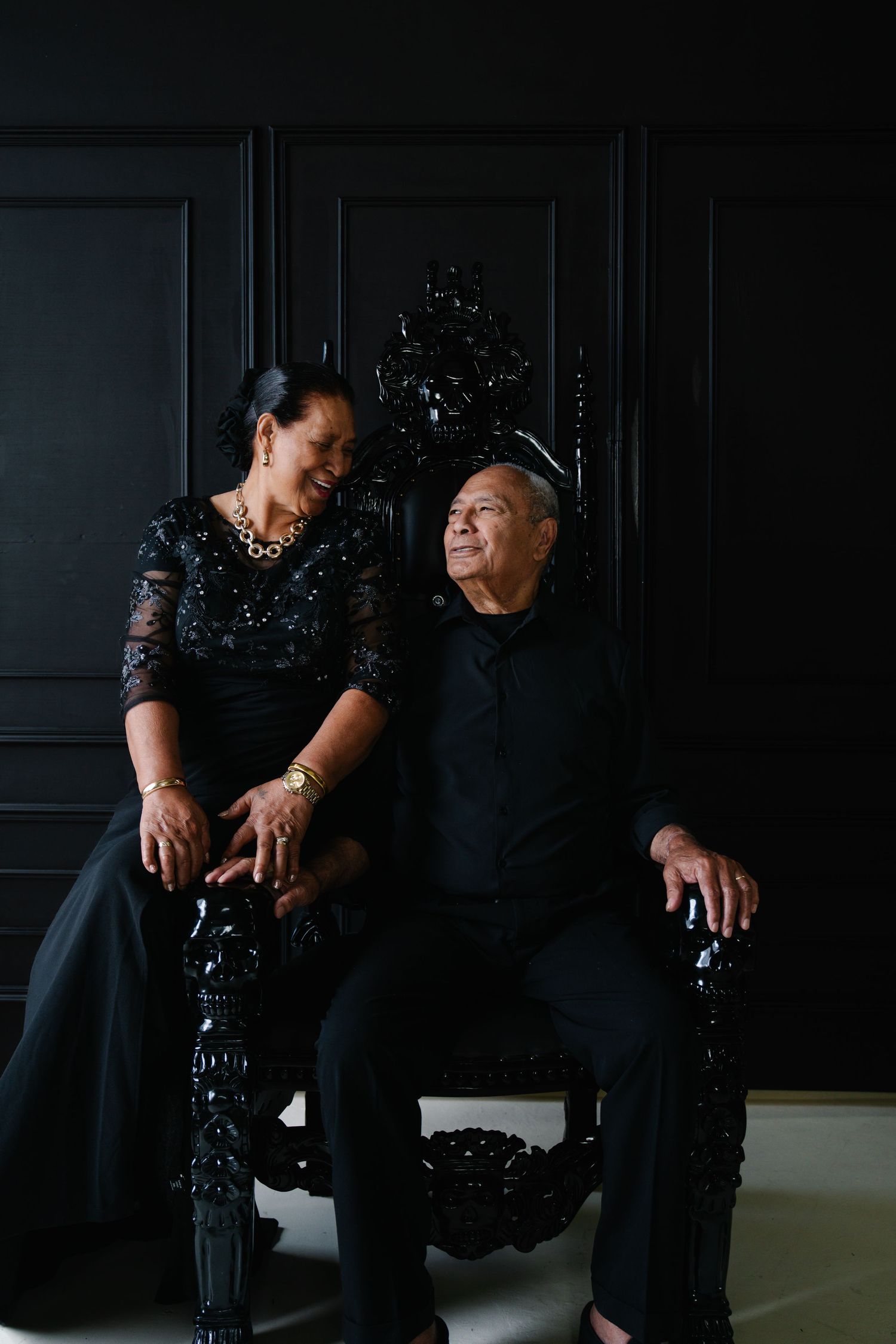 Elegant couple in matching black outfits seated on ornate black throne chair against dark wall.