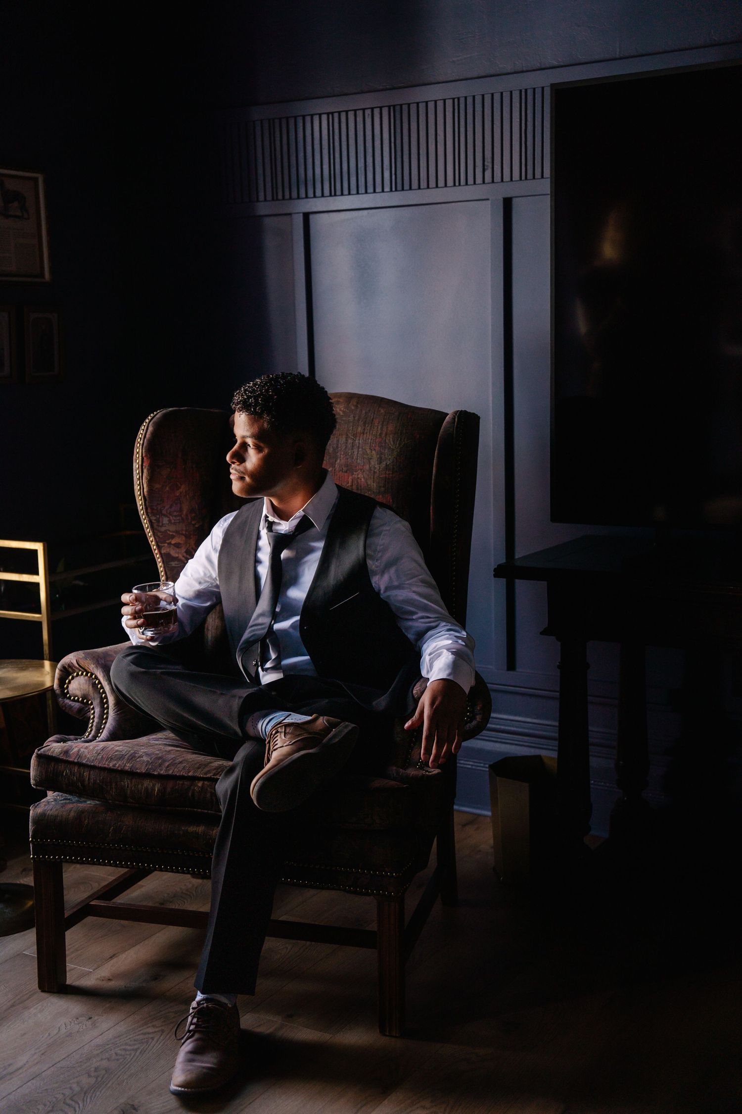Elegant man in vest and tie sitting in vintage armchair holding a drink in a dark sophisticated room.
