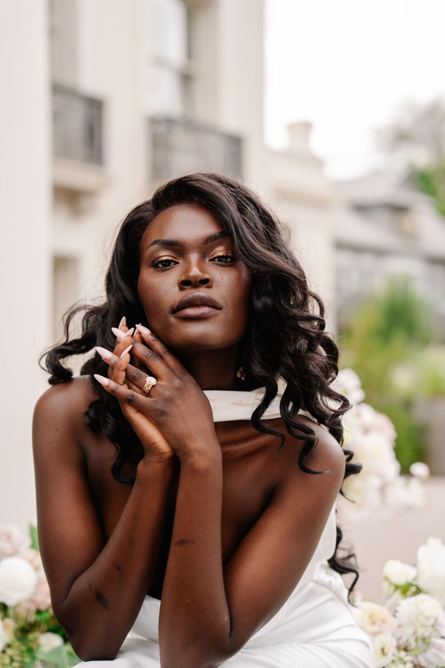 Elegant Black bride in white strapless gown poses gracefully with hands near face, showcasing curly hair and floral backdrop.