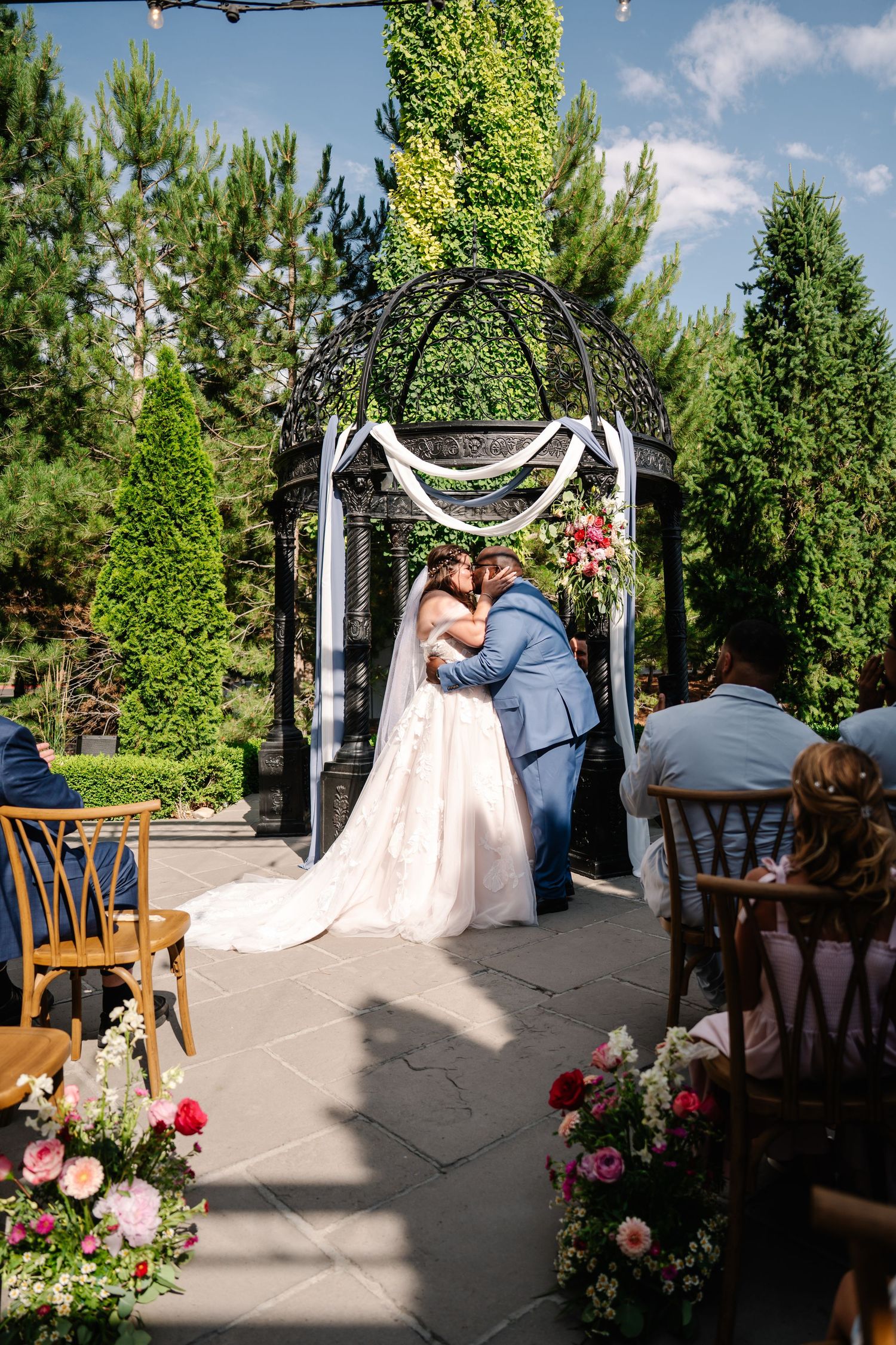 Bride and groom share first kiss under elegant iron gazebo adorned with white draping and pink flowers at outdoor wedding.