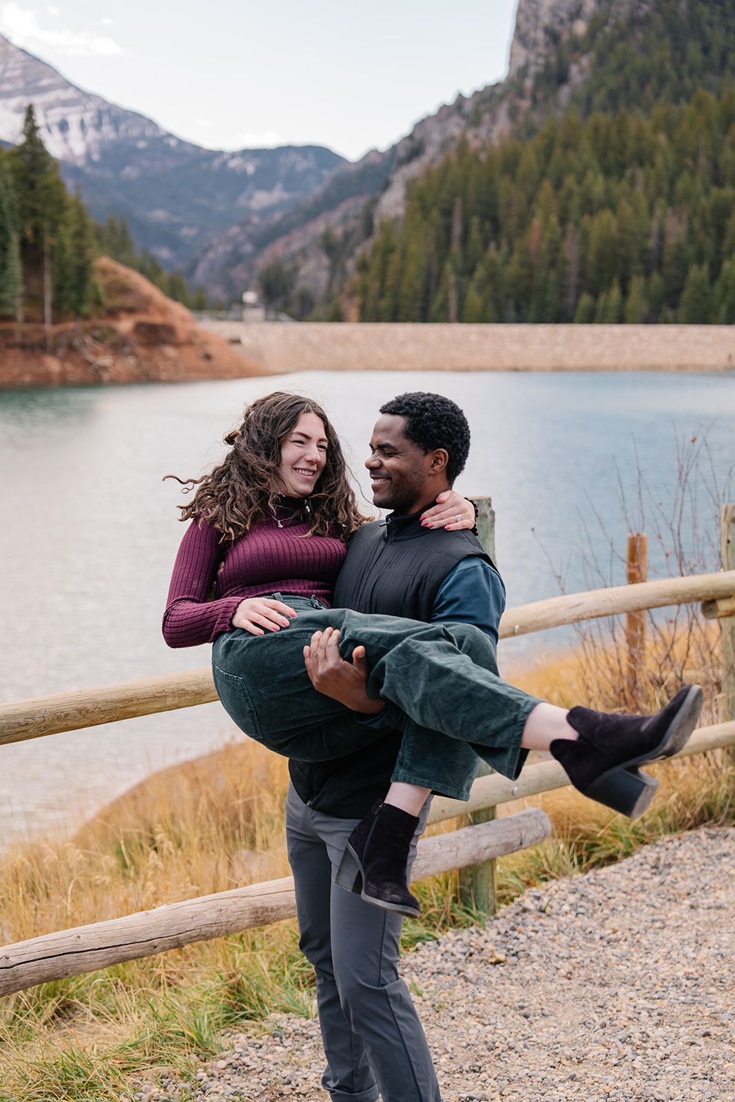 A couple shares a playful moment by a mountain lake with evergreen forests in the background.