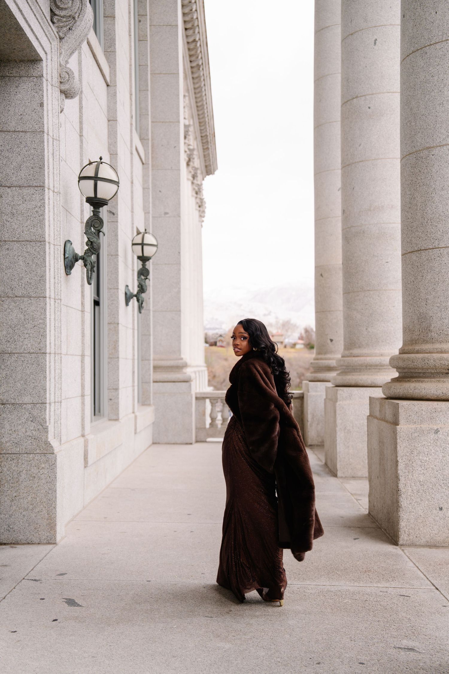Woman in flowing brown gown poses gracefully between stone columns of a grand neoclassical building.