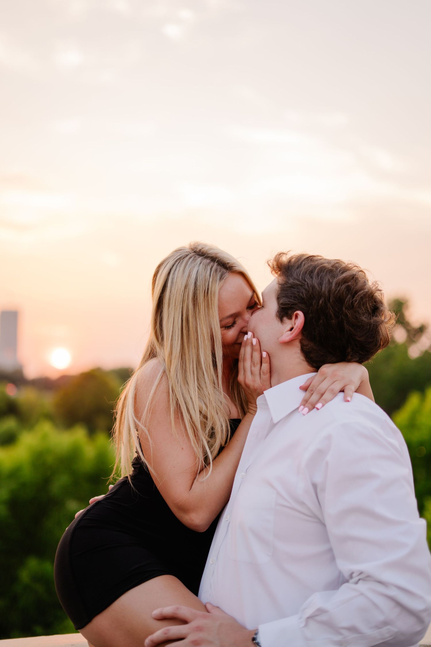 Couple sharing a romantic kiss at sunset with a warm golden sky and lush green trees in the background.