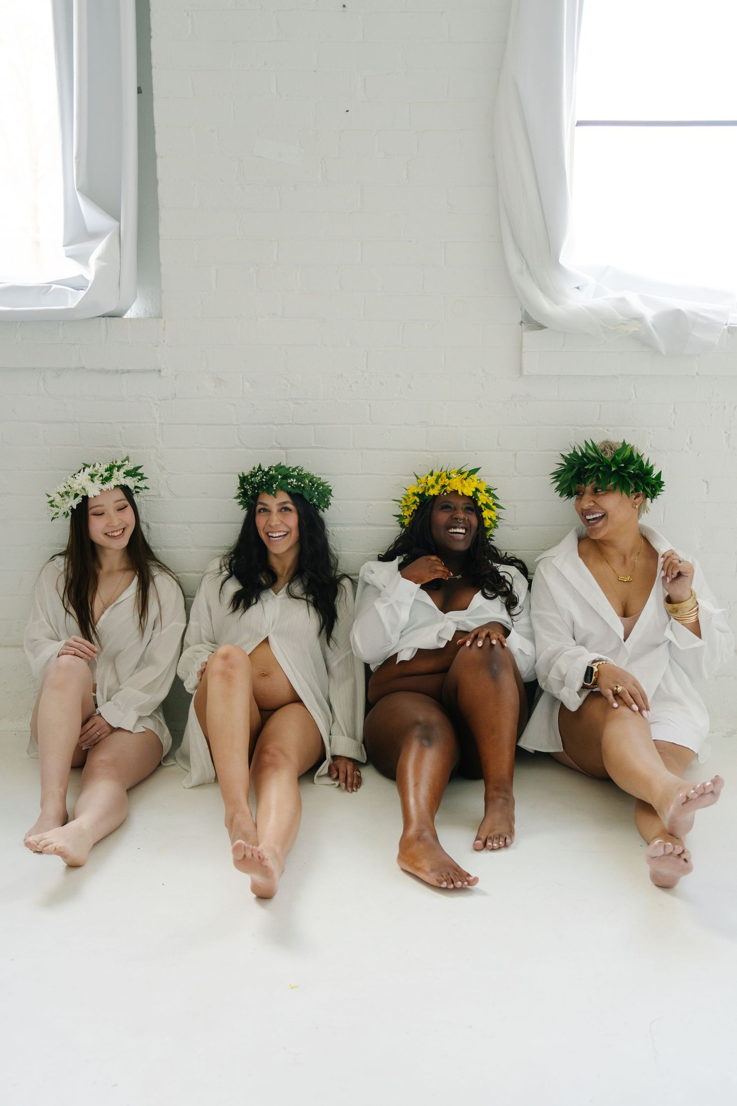 Four diverse women wearing floral crowns and white shirts sit together laughing against a white brick wall.