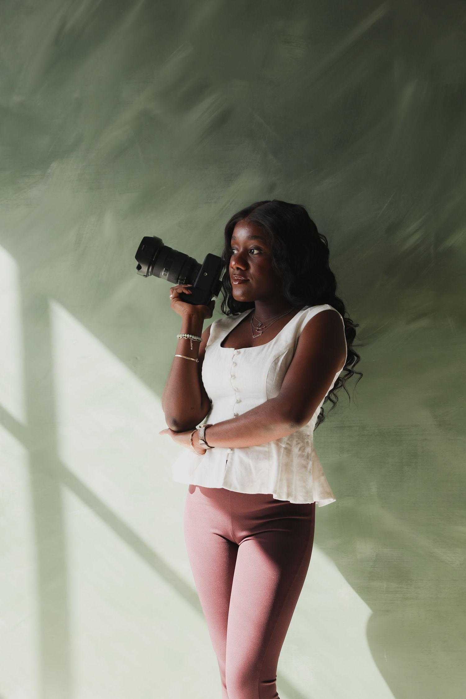 Young Black woman holding a camera, wearing white top and pink leggings, standing against a green wall in natural light.