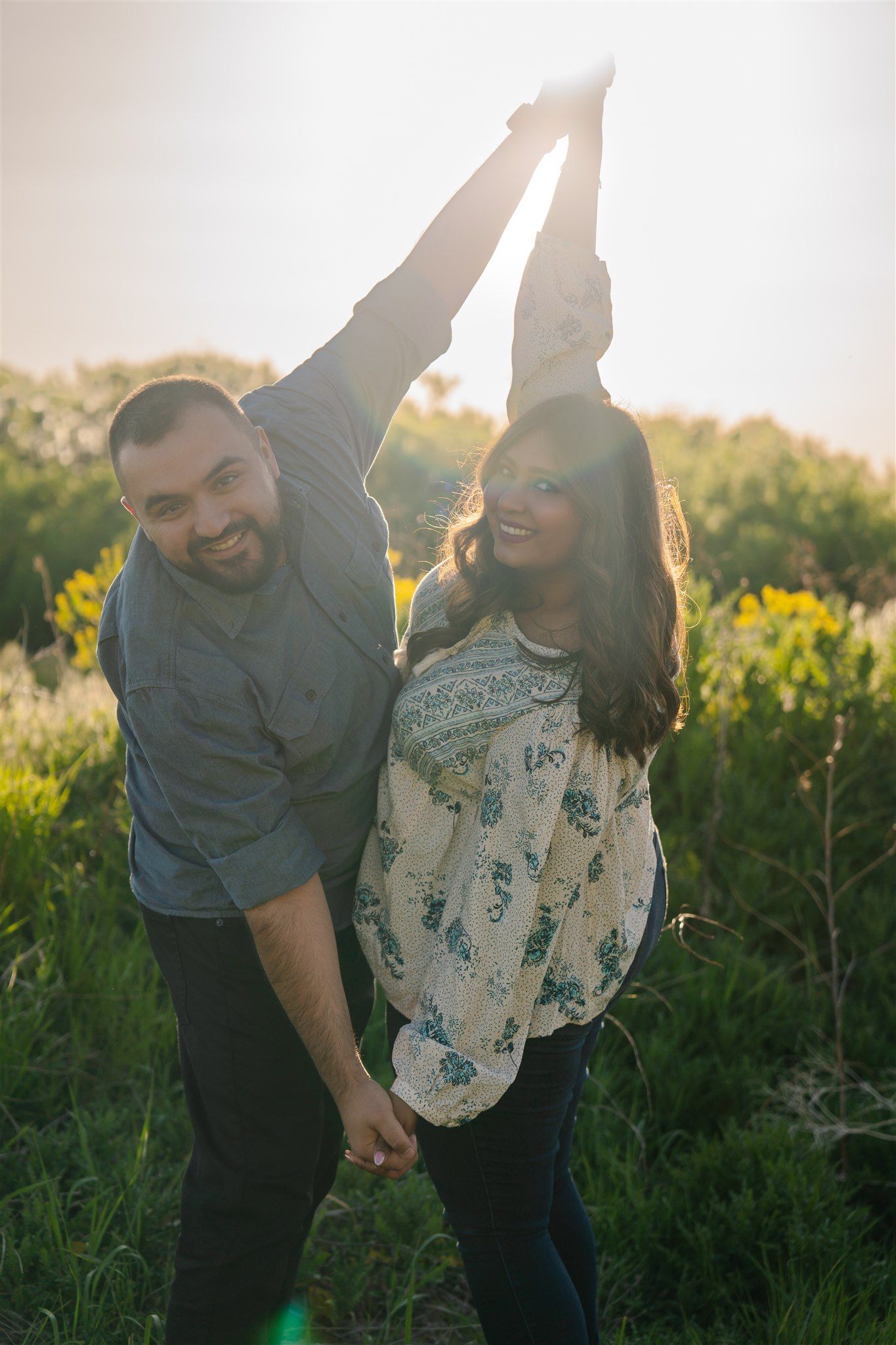 Playful outdoor couple photoshoot in golden sunlight among tall grass and wildflowers.