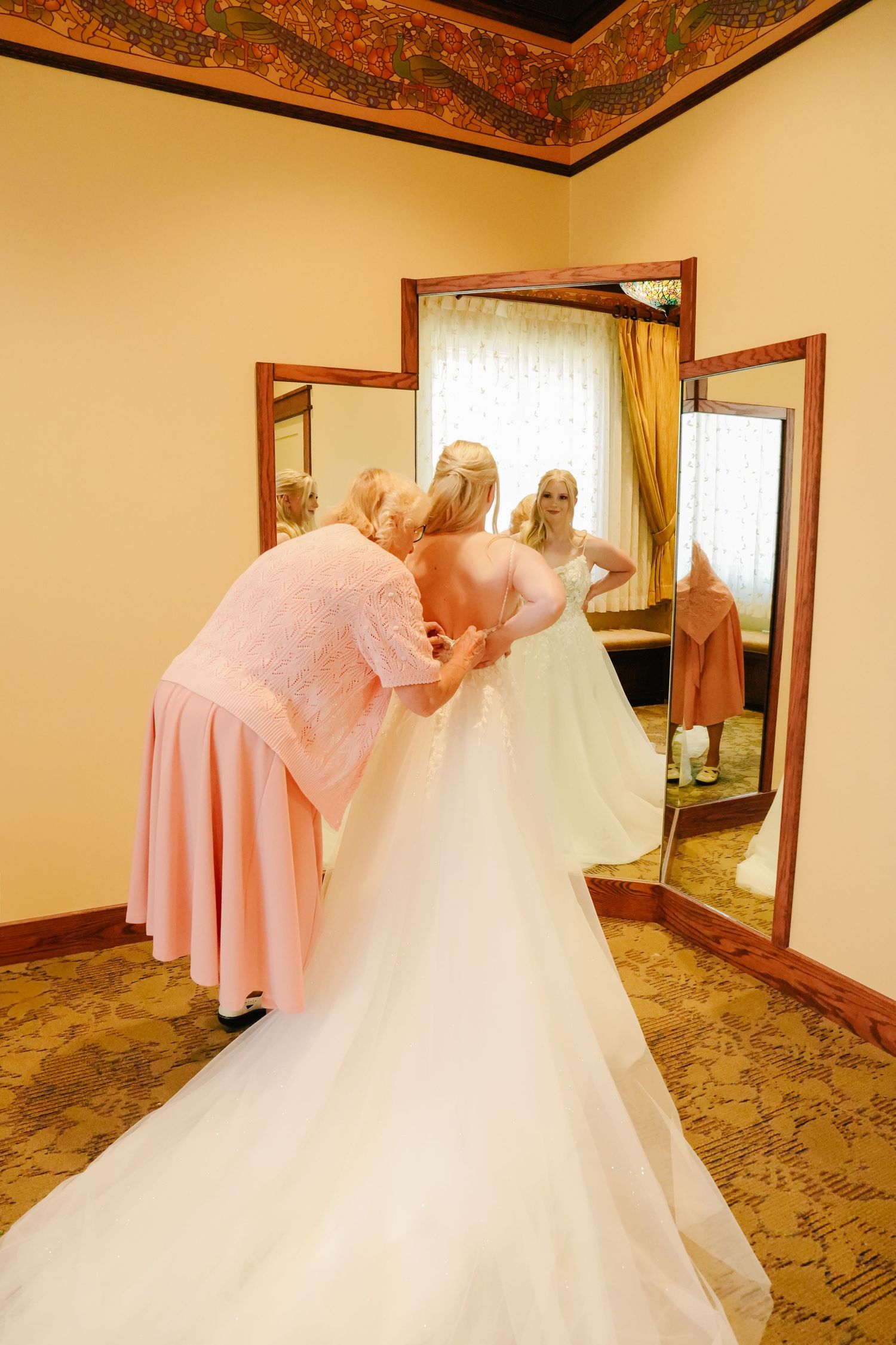 Wedding dress and pink bridesmaid dresses hanging in a hotel room with ornate ceiling details.