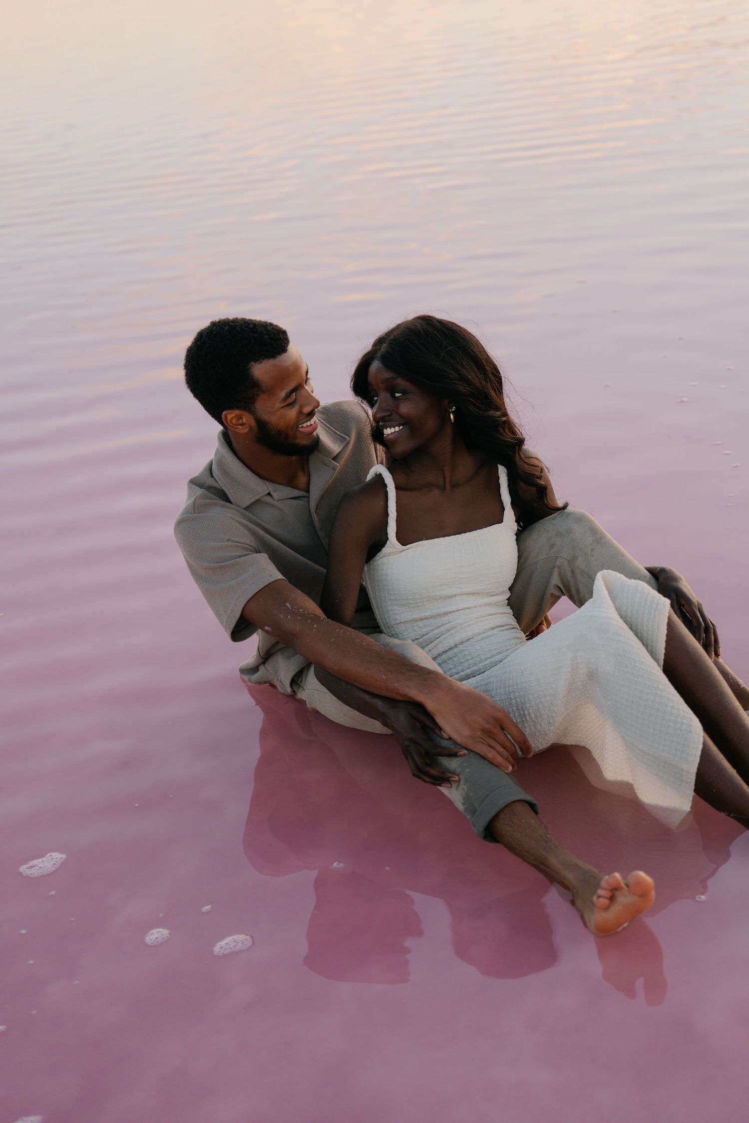 A couple shares an intimate moment while lying in shallow pink waters at sunset.