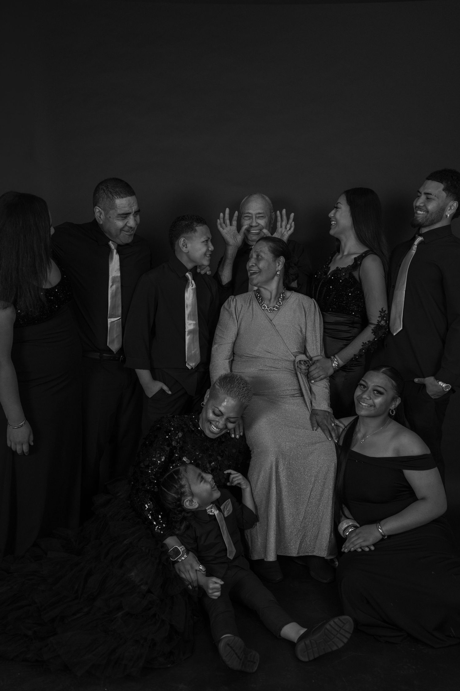 Black and white family portrait of a joyful multigenerational group posing together in formal attire against a dark background.