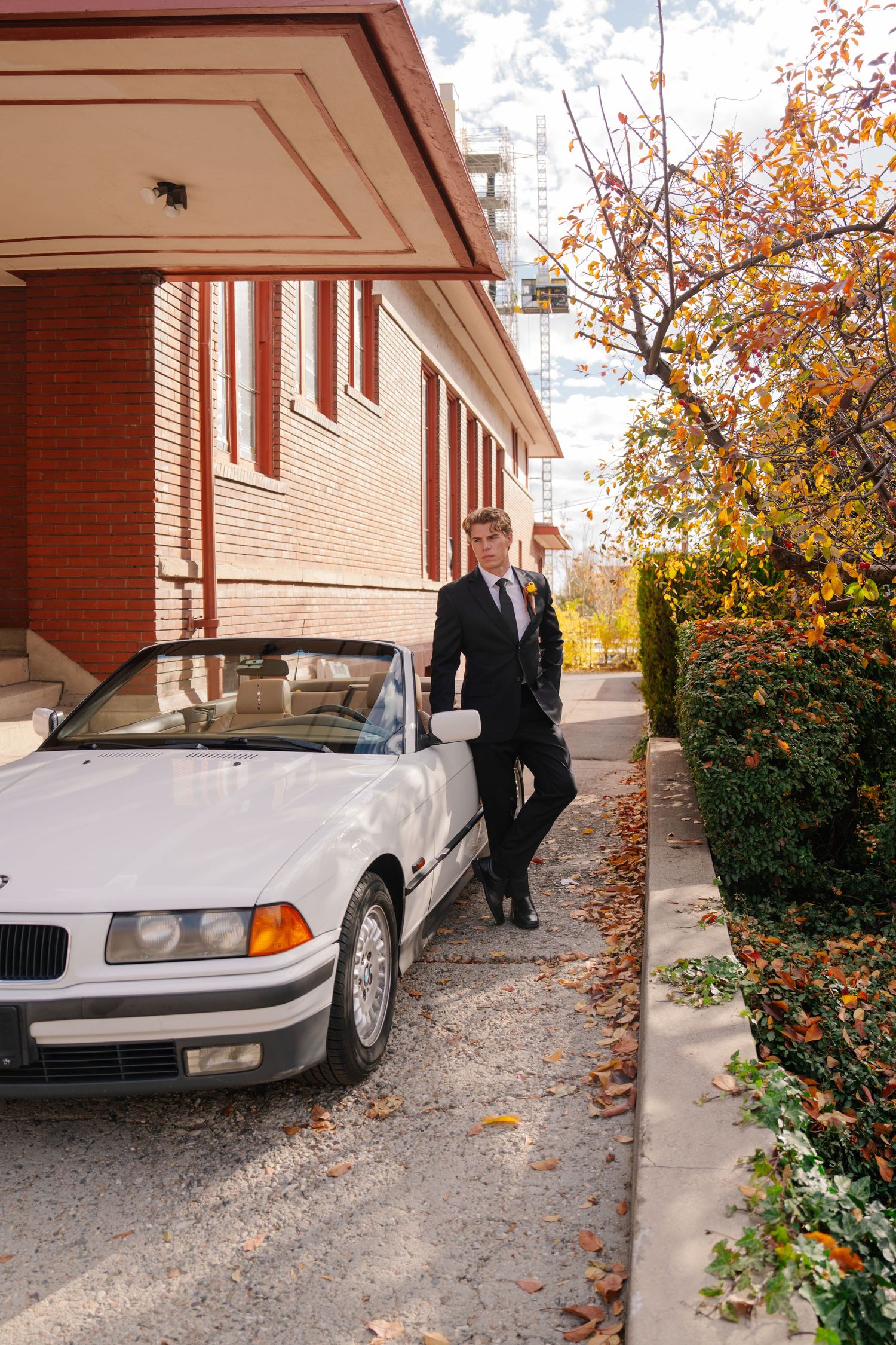 Young man in suit leaning against white BMW convertible near brick building with autumn foliage.