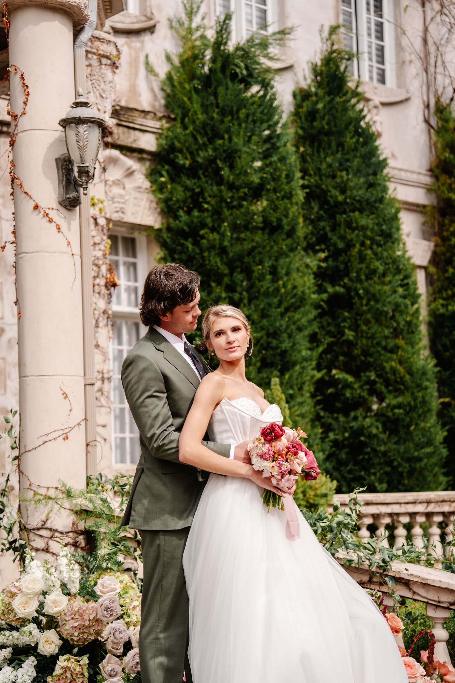 Romantic wedding couple embraces on mansion terrace surrounded by stone columns and gardens.