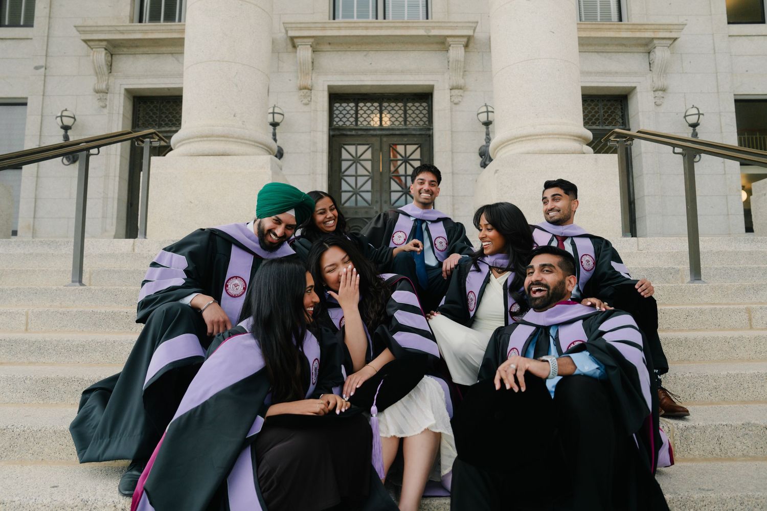 Group of university graduates in black and purple gowns celebrating on steps of a grand building.