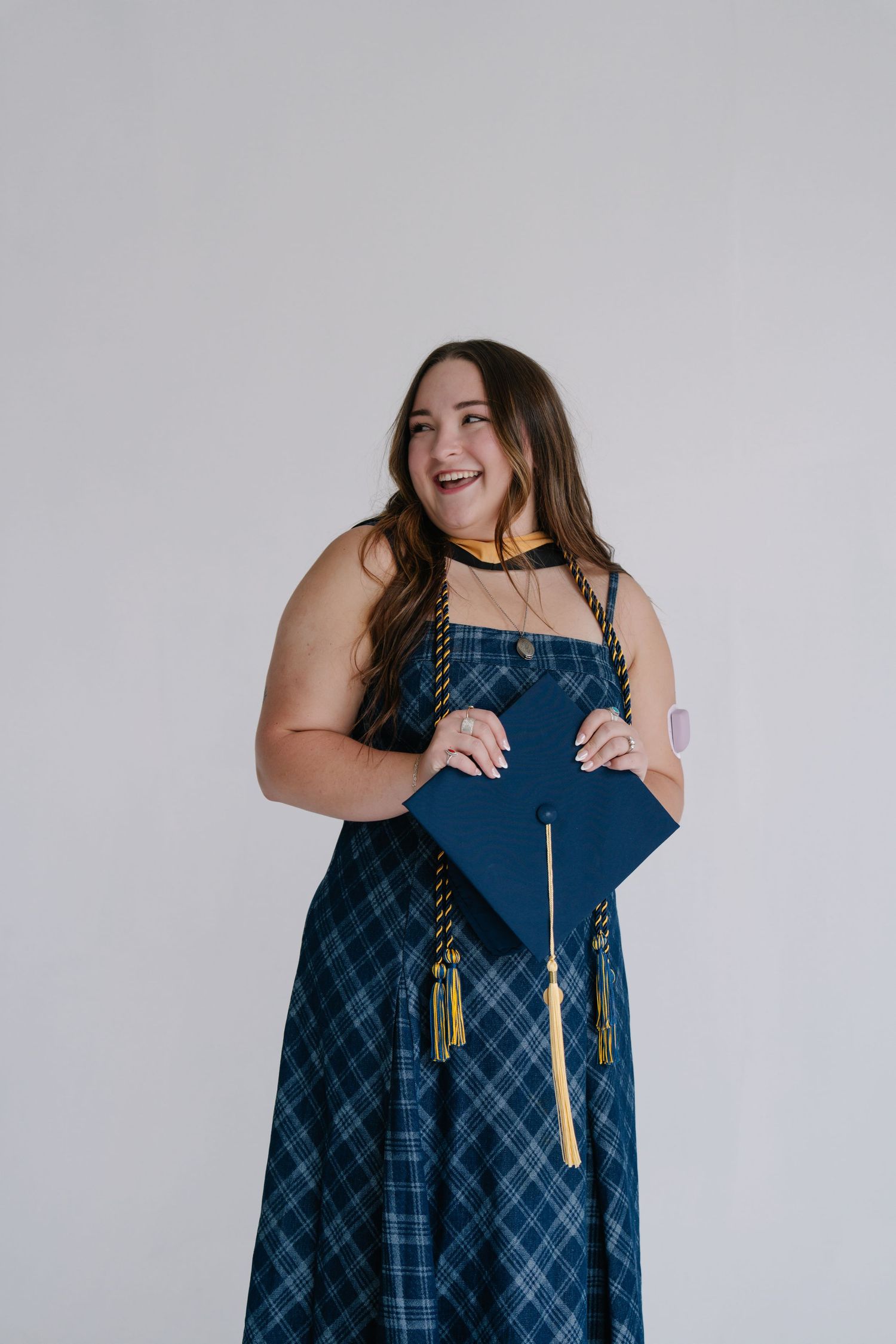 Smiling young woman in blue plaid dress holding navy graduation cap with gold tassel, celebrating academic achievement.
