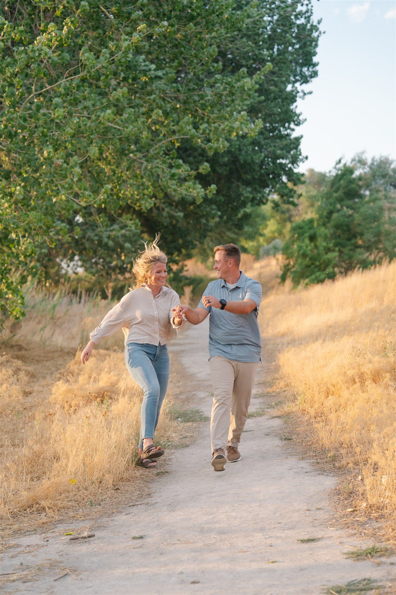 Couple runs playfully together on a dirt path surrounded by golden grass and green trees.