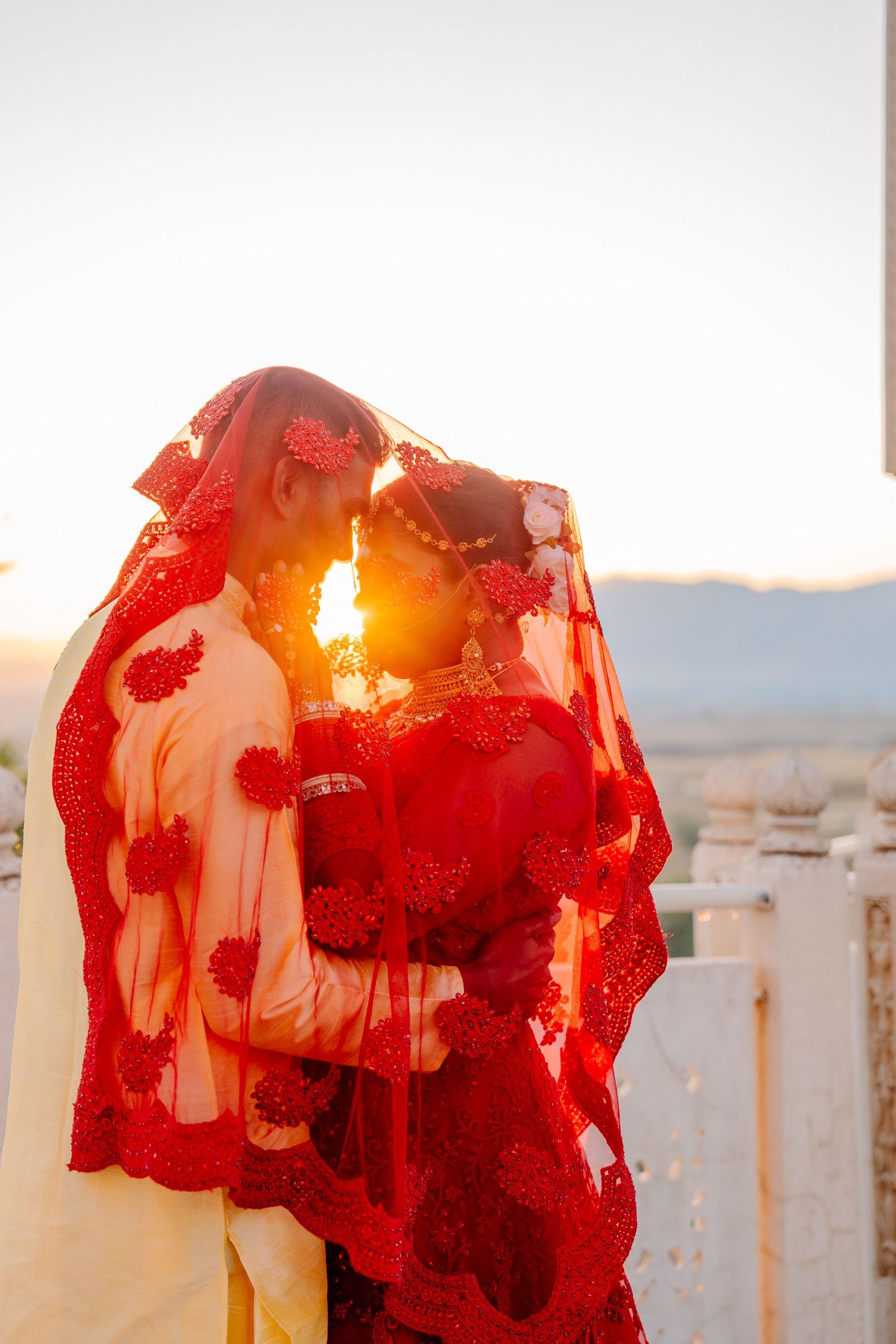 A couple embracing under a red dupatta at sunset creating a romantic silhouette.