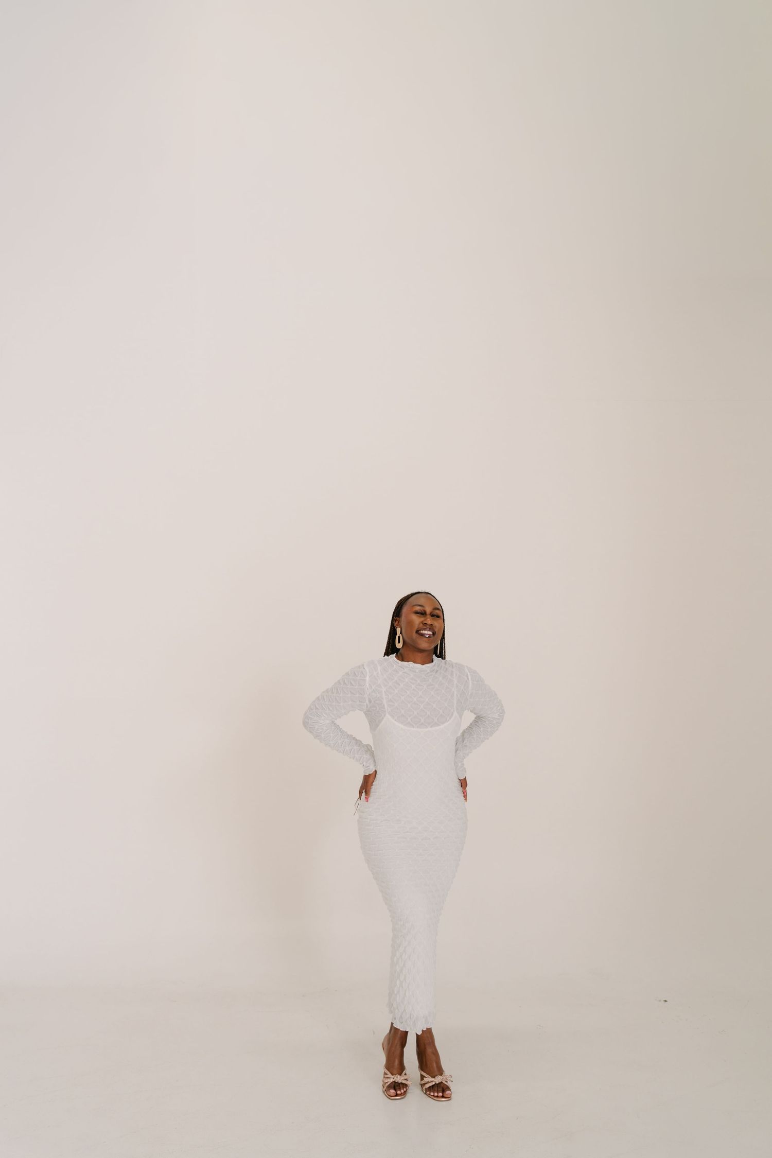 A smiling Black woman poses confidently in an all-white outfit with heels against a white background.