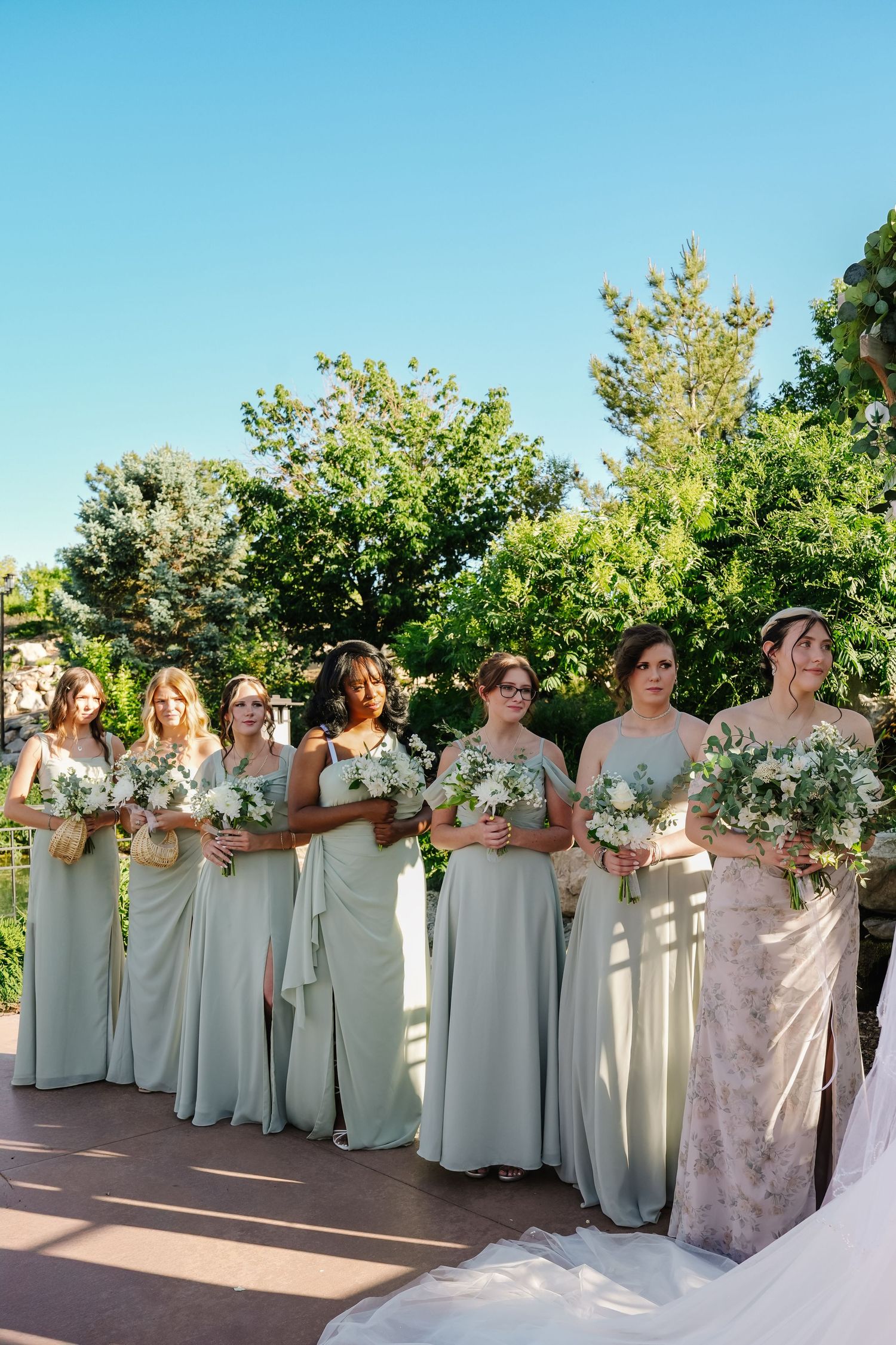 Bridesmaids in sage green dresses pose together holding white floral bouquets in an outdoor garden setting.