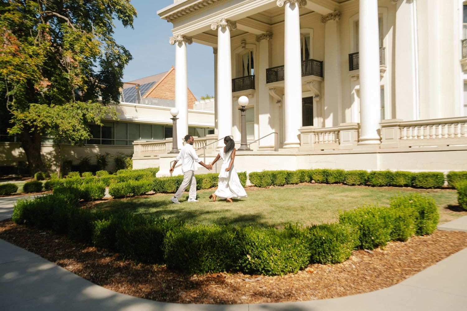 Couple in white outfits holding hands on lush green lawn outside elegant white neoclassical building with tall columns.