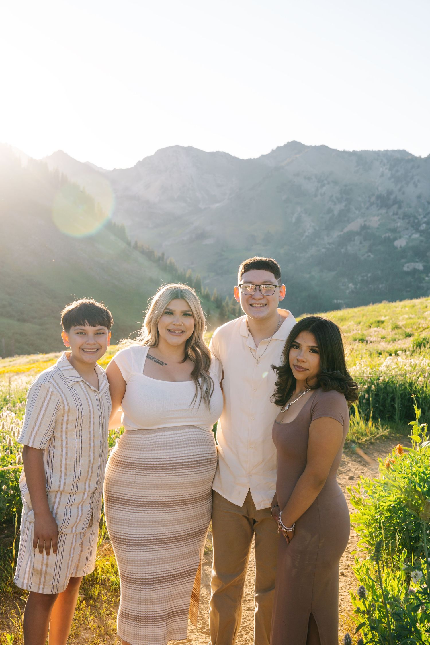 Four people smiling together in a sunny mountain meadow with green hills and peaks in the background.