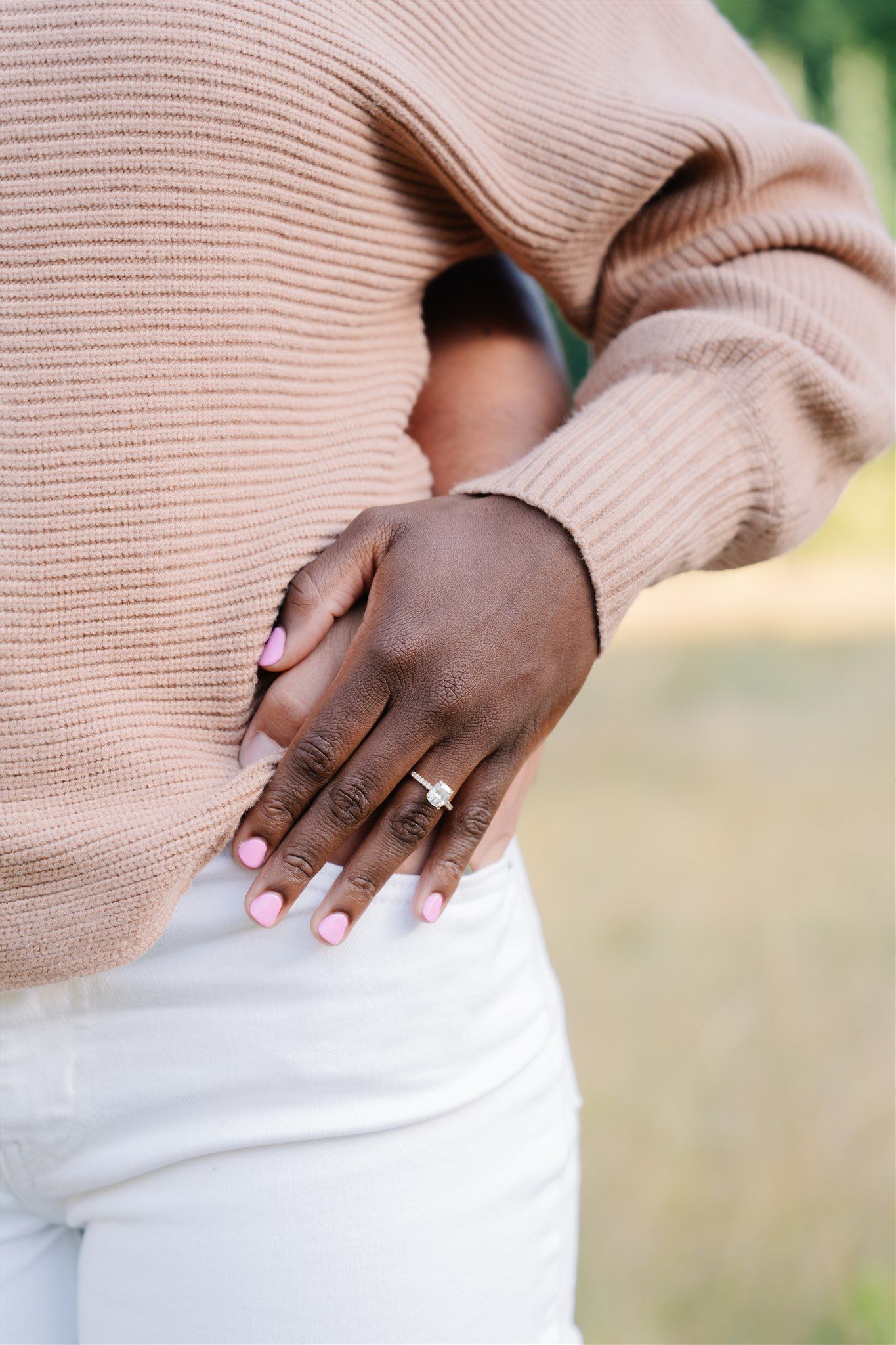 Close-up of hands wearing a pink sweater and engagement ring against a light background.
