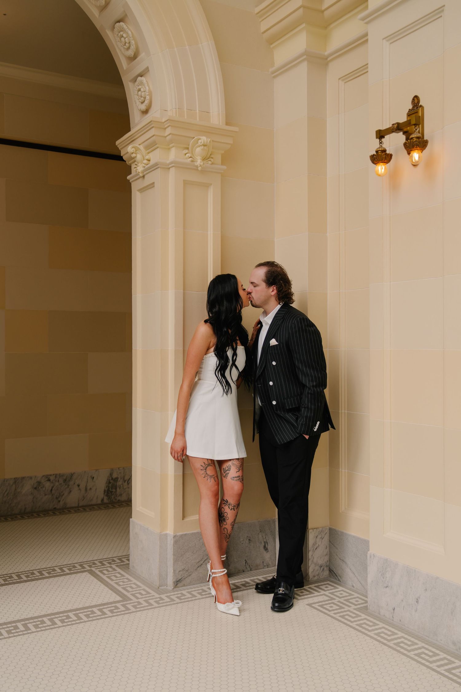 Couple sharing a kiss in elegant courthouse hallway, bride in white mini dress with tattoos, groom in pinstripe suit.