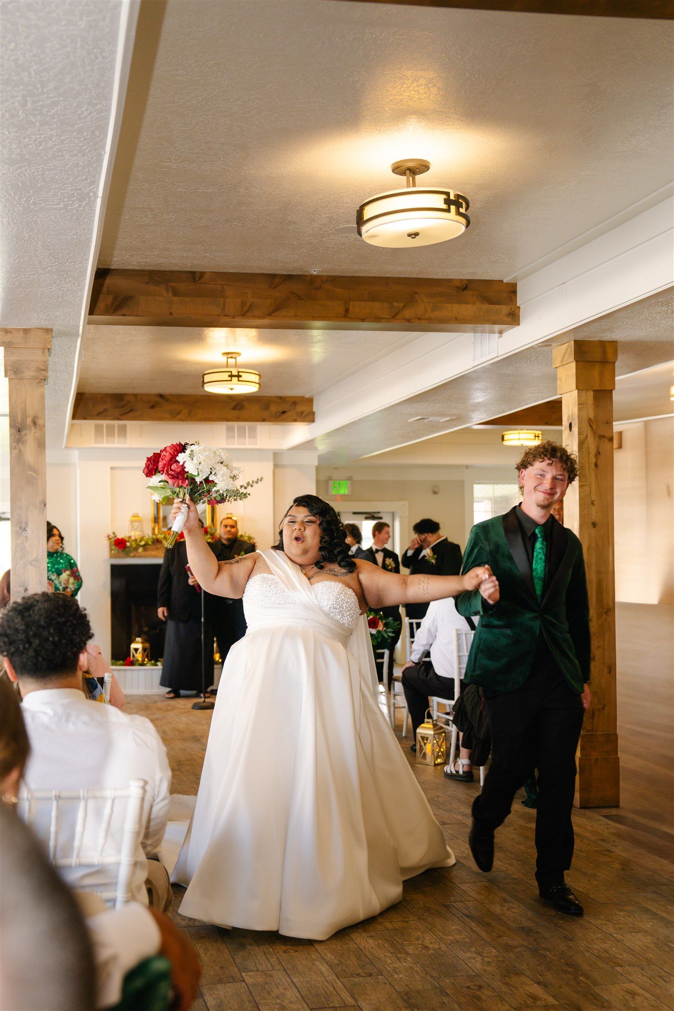 Wedding couple dances joyfully in a rustic venue with wooden beams and ceiling lights.