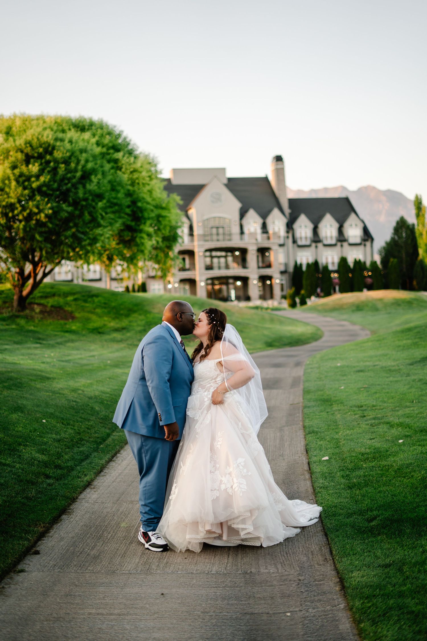 A wedding couple shares an intimate moment on a winding path leading to a grand estate building.