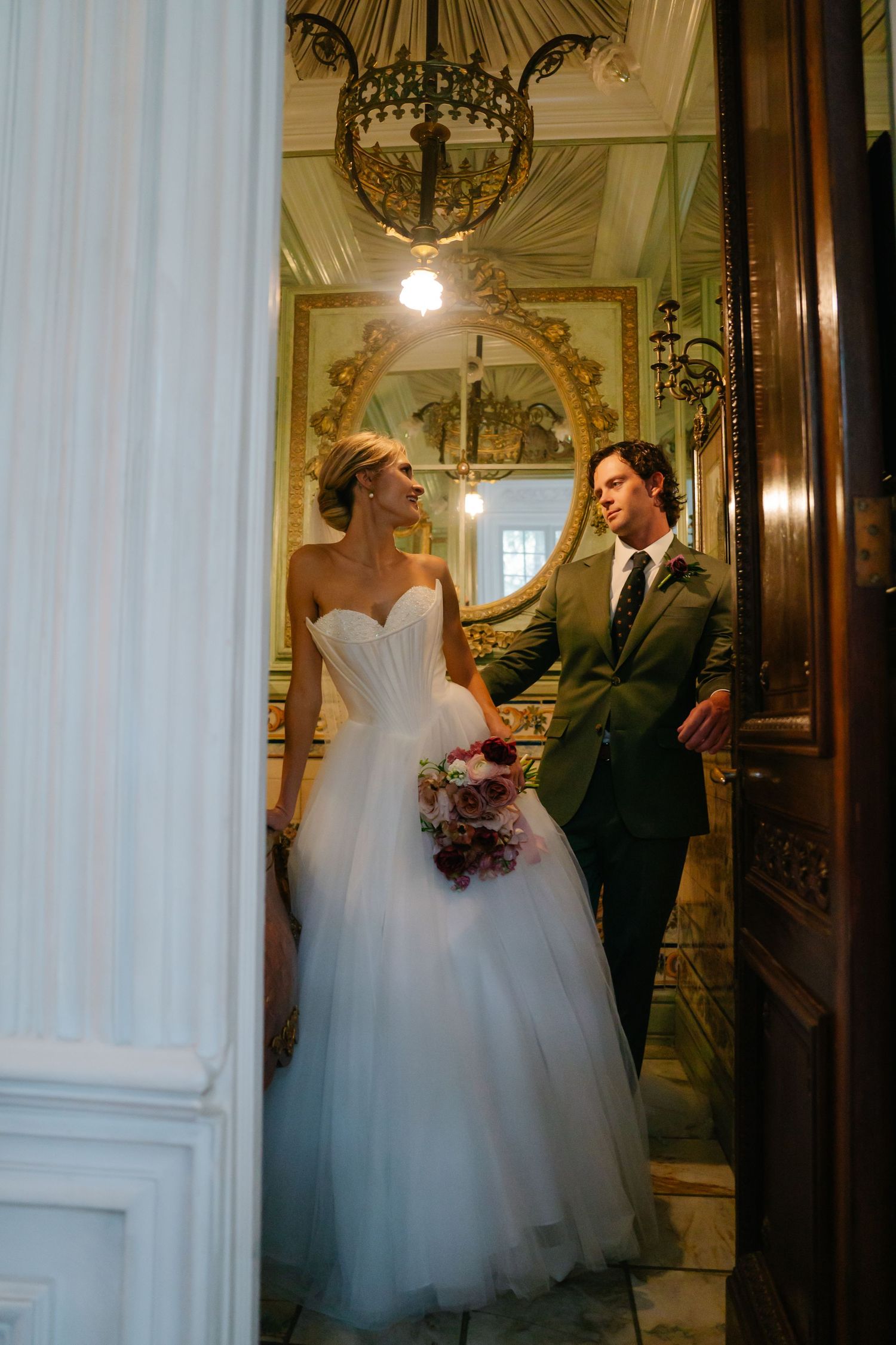 Couple sharing a joyful moment in an elegant hallway with chandelier and ornate architectural details.