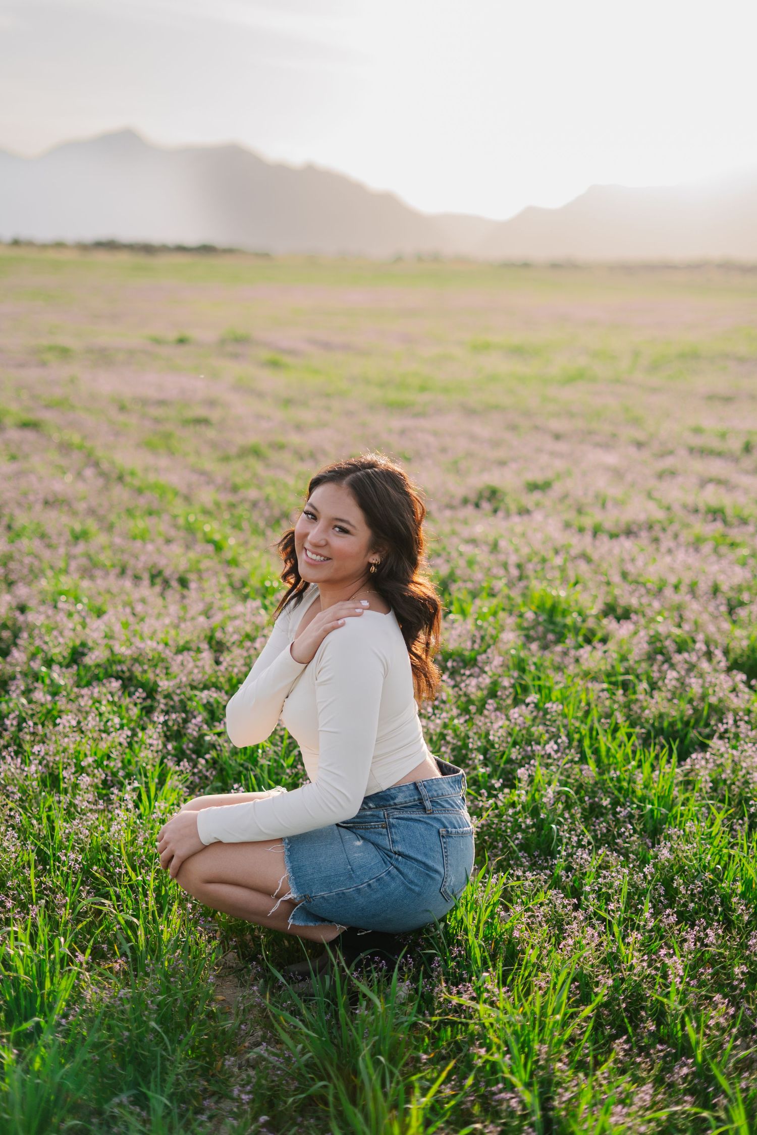 Young woman in white top and denim skirt crouching in a blooming field with mountains at sunset.