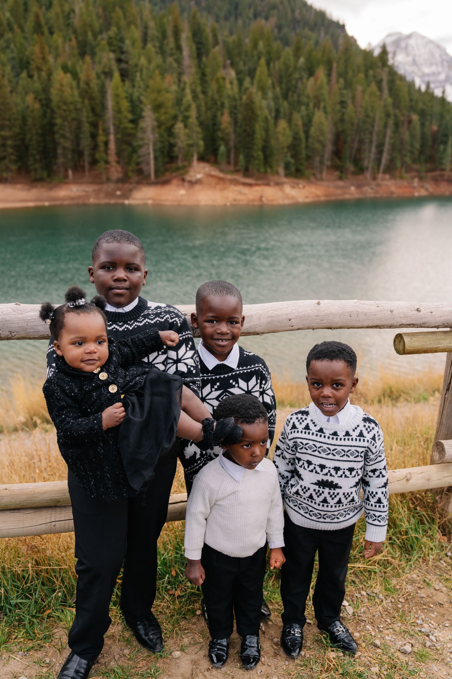 Five Black children in matching Nordic sweaters pose by a turquoise mountain lake surrounded by evergreen trees.