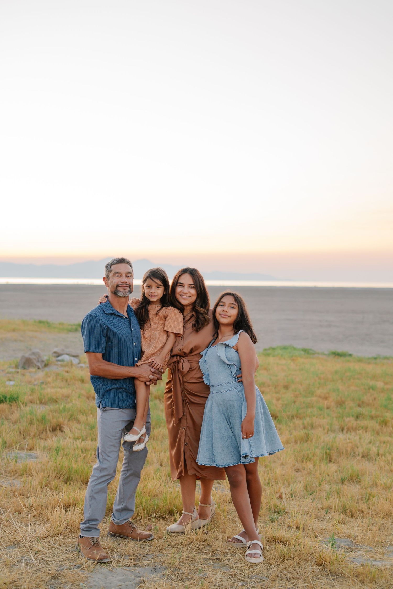 Family of four posing at sunset near a coastal field, parents and two daughters smiling in warm golden light.
