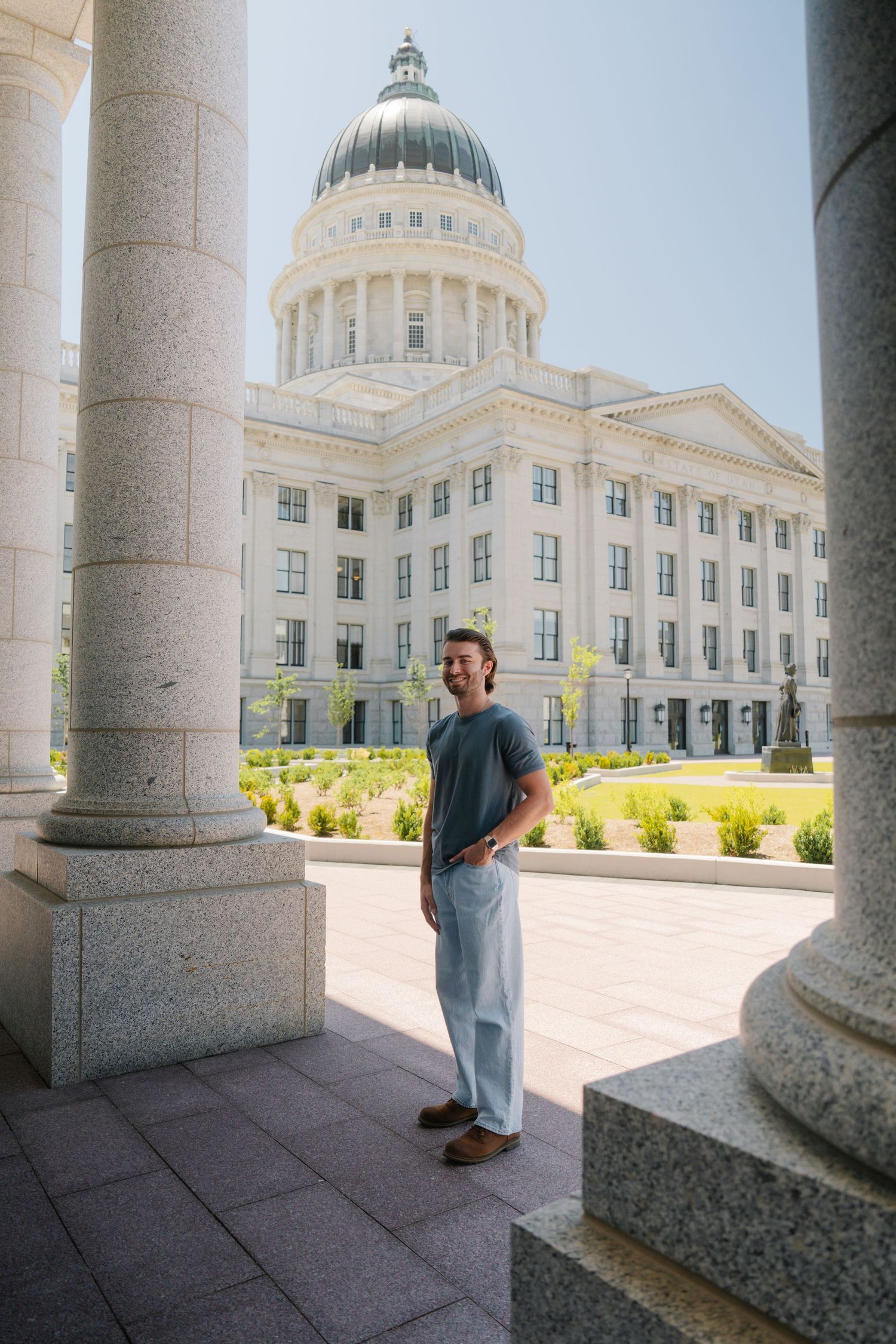 Man smiling in front of Utah State Capitol building, standing between granite columns on a sunny day.