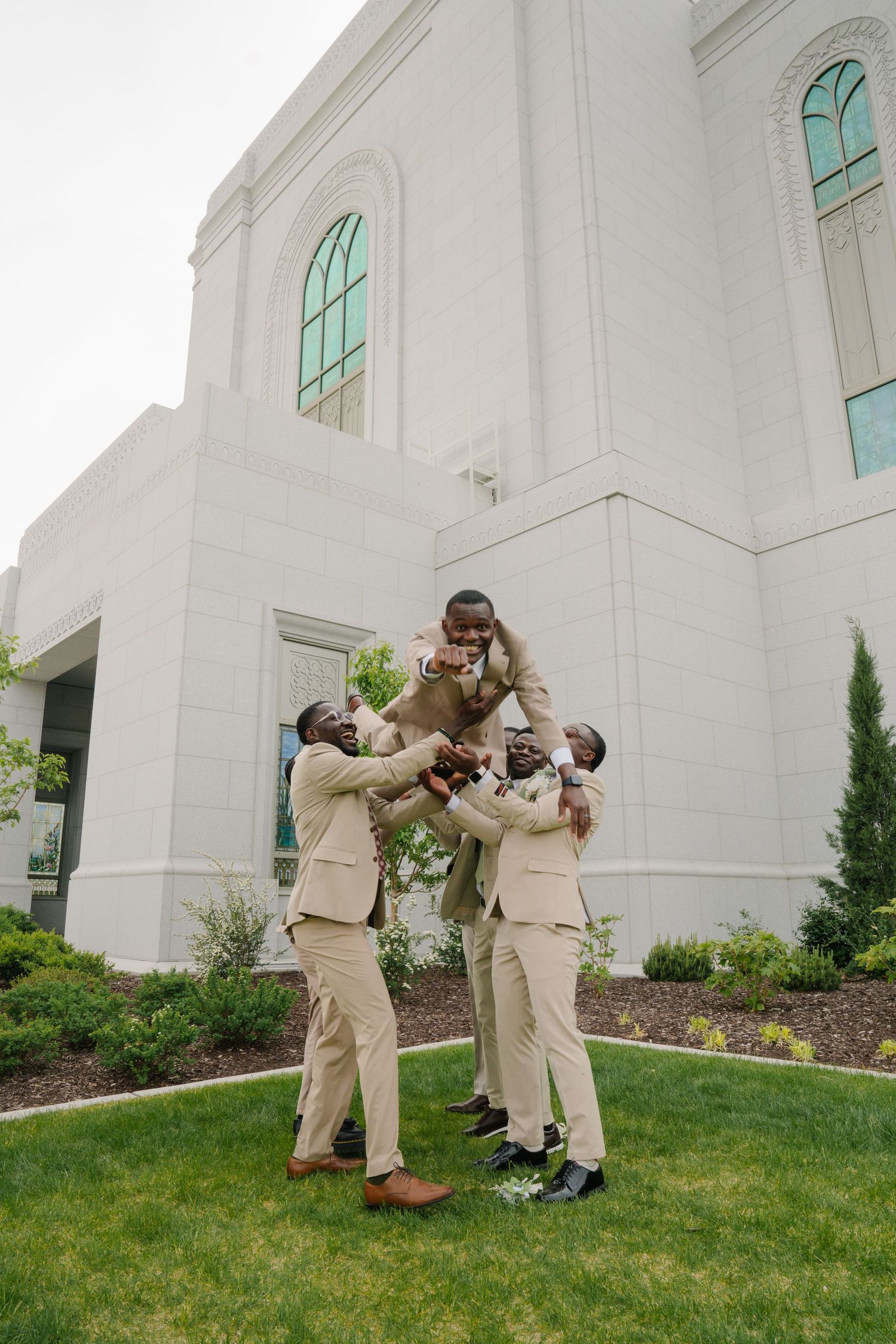 Groomsmen in tan suits lifting the groom outside a white wedding venue on a green lawn.