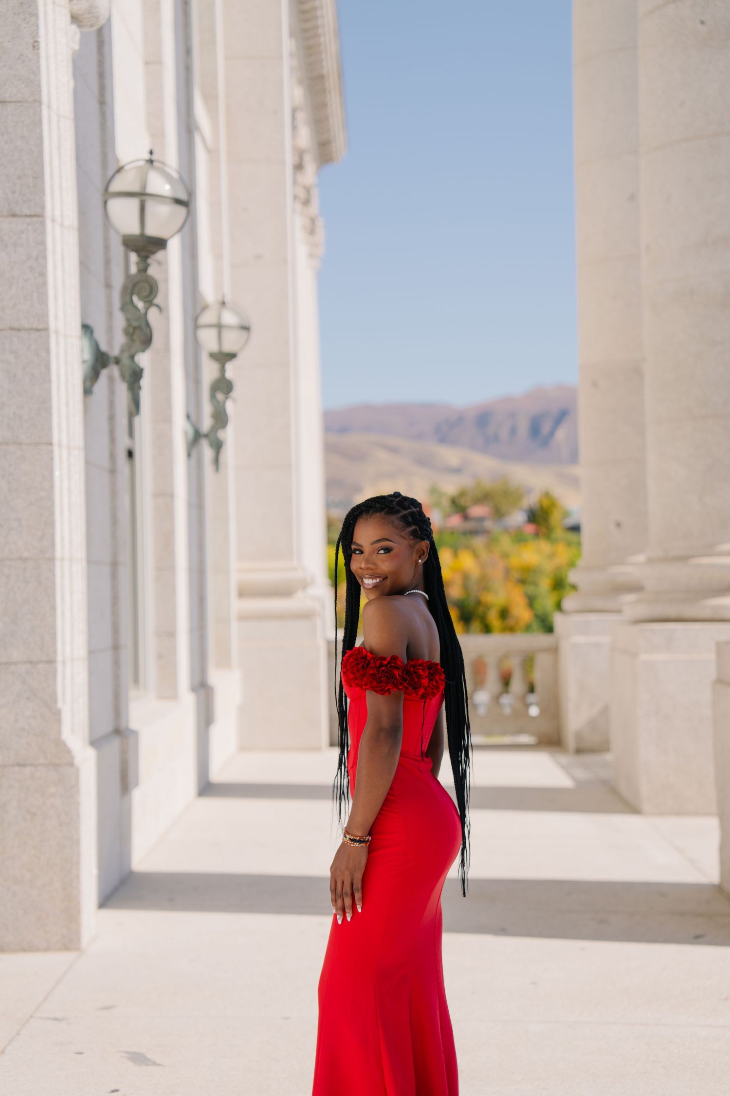 Young Black woman in elegant red off-shoulder gown poses gracefully between white columns with mountain views behind her.