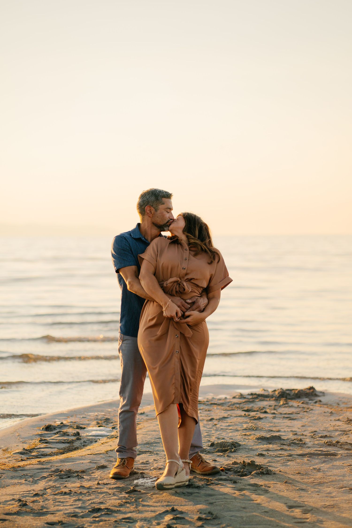 Couple sharing a romantic kiss on a sandy beach at golden hour sunset by the ocean.