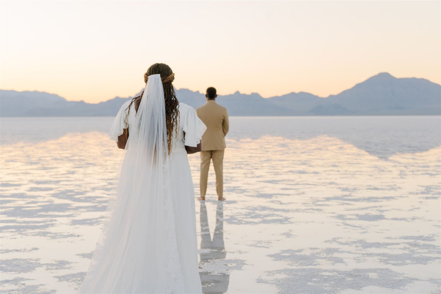 A couple walks along a salt flat during sunset with mountains in the distance.